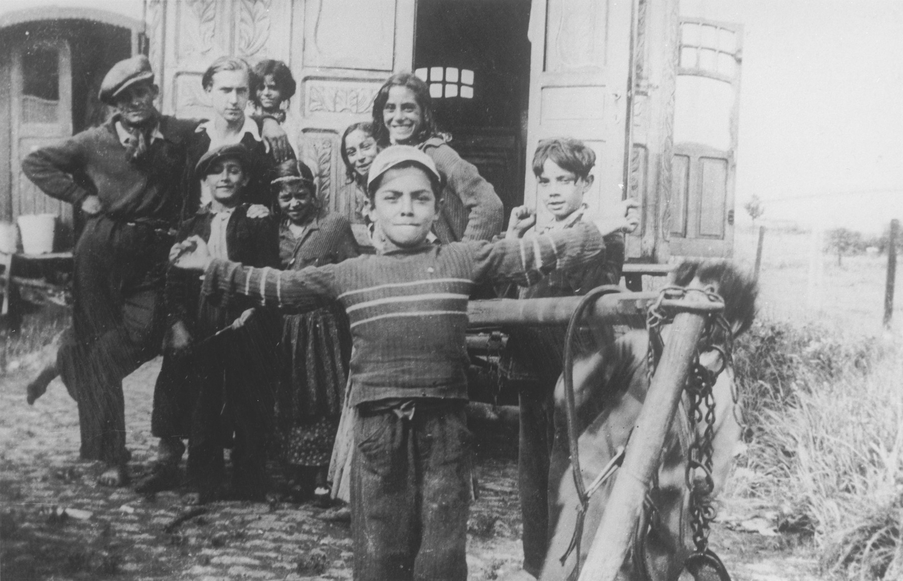 A Romani child poses in front of a caravan with his arms outstretched, while his family looks on.

Pictured in the back, second from the left, is Jan Yoors.