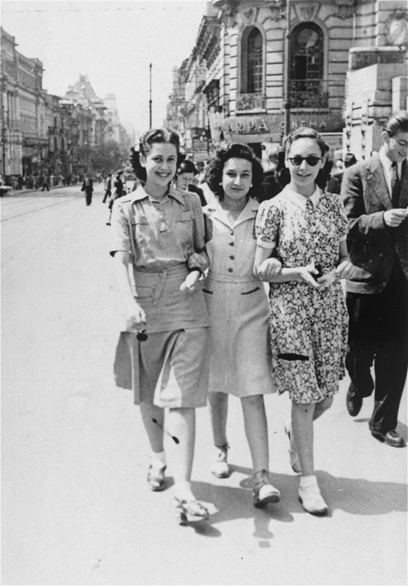Three Jewish women stroll down a commercial street in Bucharest.  

Pictured from left to right are: Valerie Zigmund (Cernes), Jeanine Gutman, and Anna Erlic.