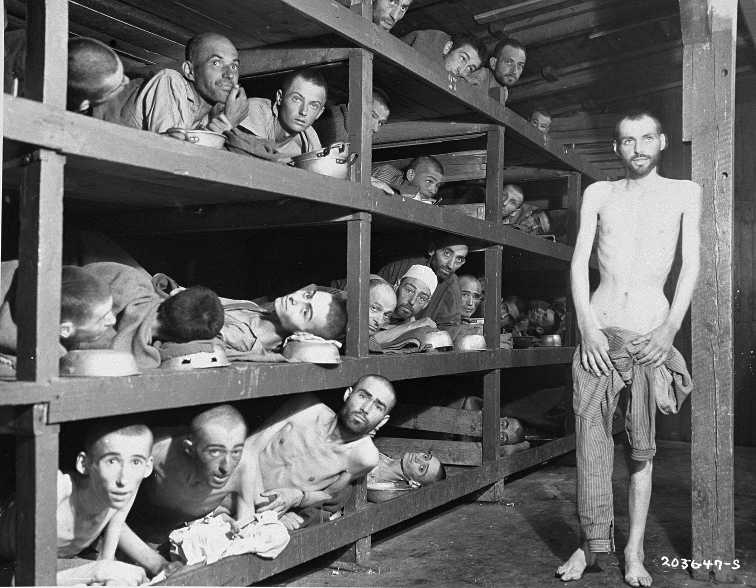Former prisoners of the "little camp" in Buchenwald stare out from the wooden bunks in which they slept three to a "bed."  

Elie Wiesel is pictured in the second row of bunks, seventh from the left, next to the vertical beam.  

The man in the bottom left hand corner has been identified as Michael Nikolas Gruner, originally from Hungary, Gershon Blonder Kleinman or Yosef Reich.  Isaac Reich is in the bottom row, second from the right and Max Hamburger is on the bottom row, fourth from the left.  Perry Shulman from Klimitov, Poland is on the top bunk, second from the left (looking up).  The man in the second row, third from left has been identified as Dawid Najman.  The man in the second row, fourth from the left has been identified as Abraham Hipler;  Berek Rosencajg from Lodz or Zoltan Gergely from Cluj.  The man on the third bunk from the bottom, third from the left, has been identified alternatively as Ignacz (Isaac) Berkovicz, Abraham Baruch and Joseph Icovic )from Tecovo) .  Juraj (now Naftali) Furst is pictured in the third bunk, fifth from the left.  The man standing on the right has been identified as Chaim David Halberstam, and also as Simon Toncman.  In the top left bunk, far right is Oscar Kleinshpitz (later Haberkorn).  The man pictured in the middle bunk, fifth from the left (wearing a white cap) has been identified as Simon Brommet,.  Simon was born in Amsterdam and was captured in Belgium. He survived a labor camp in Auschwitz, was marched to Buchenwald in the winter of 1945, and was liberated by American troops on April 11, 1945.

Original caption reads:  "Slave laborers  pedred from their tomb-like, wooden bunks at the concentration camp at Buchenwald, near Weimar, Germany, when U.S. troops liberated the area.  Approximately 21,000 inmates - Russian, Polish, French, Italian, and Czechoslovakian - were freed.  They were described as "the living dead".  The camp doctors agreed that a t least 6,000 inmates had died as the result of German brutality during March, 1945.  In the Winter of 1939, the prisoners died at the rate of 900 daily.  In 10 years, more than 70,000 persons were starved and butchered in this atrocity center."