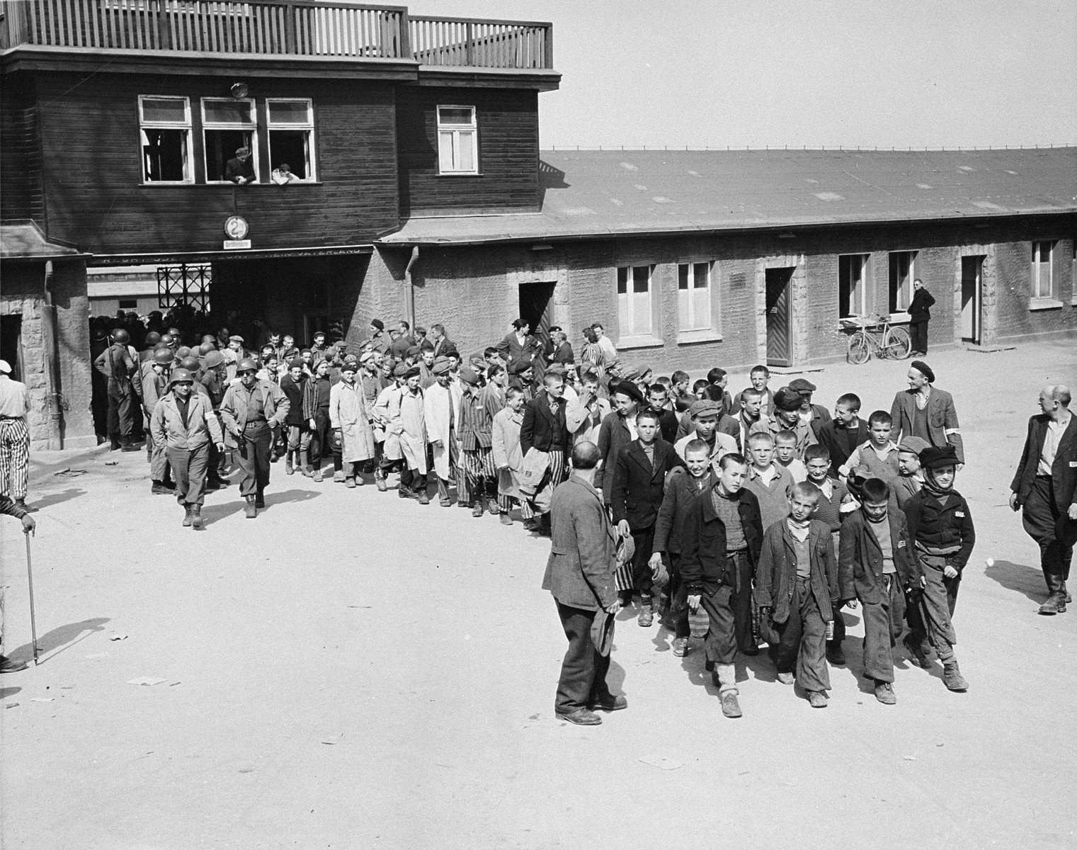 Escorted by American soldiers, a transport of child survivors of Buchenwald file out of the main gate of the camp.

The original caption reads, "Internees at the Buchenwald Concentration Camp near Weimar Germany march from the camp to receive treatment at an American hospital after the camp was captured by U.S. Third Army troops."

The boys are accompanied by American soldiers and directed by elements of the camp underground who watd over them, including Polish Jews Yakov Werber and Eli Grinbaum (on the right). 

Among the children pictured are Izio Rosenman (head of column), Jacques Finkel, Charles Finkel, Fredek Margolis, Lalek Russ, Salek Sandowski, George Goldbloom and ? Zylber.  Misho Frailich, Willi Fogel, A. Grossman, Lotci Miller, Laiza Grynberg, Usha Grynberg, David Perlmutter, Marek Lodzinsky, Yankel Kapelush, Yosel Dziubak, Loyosh Hershkovitz, ? Yakubovitz, Reuven Wekselman, Stanley Weinstein, Herschek Zeit, Henryk Kolber, Jacques Werber and Philip Kaner.  Mor Stern is the boy in the beret and white coat, with right arm hanging down straight just right of the two American soldiers.