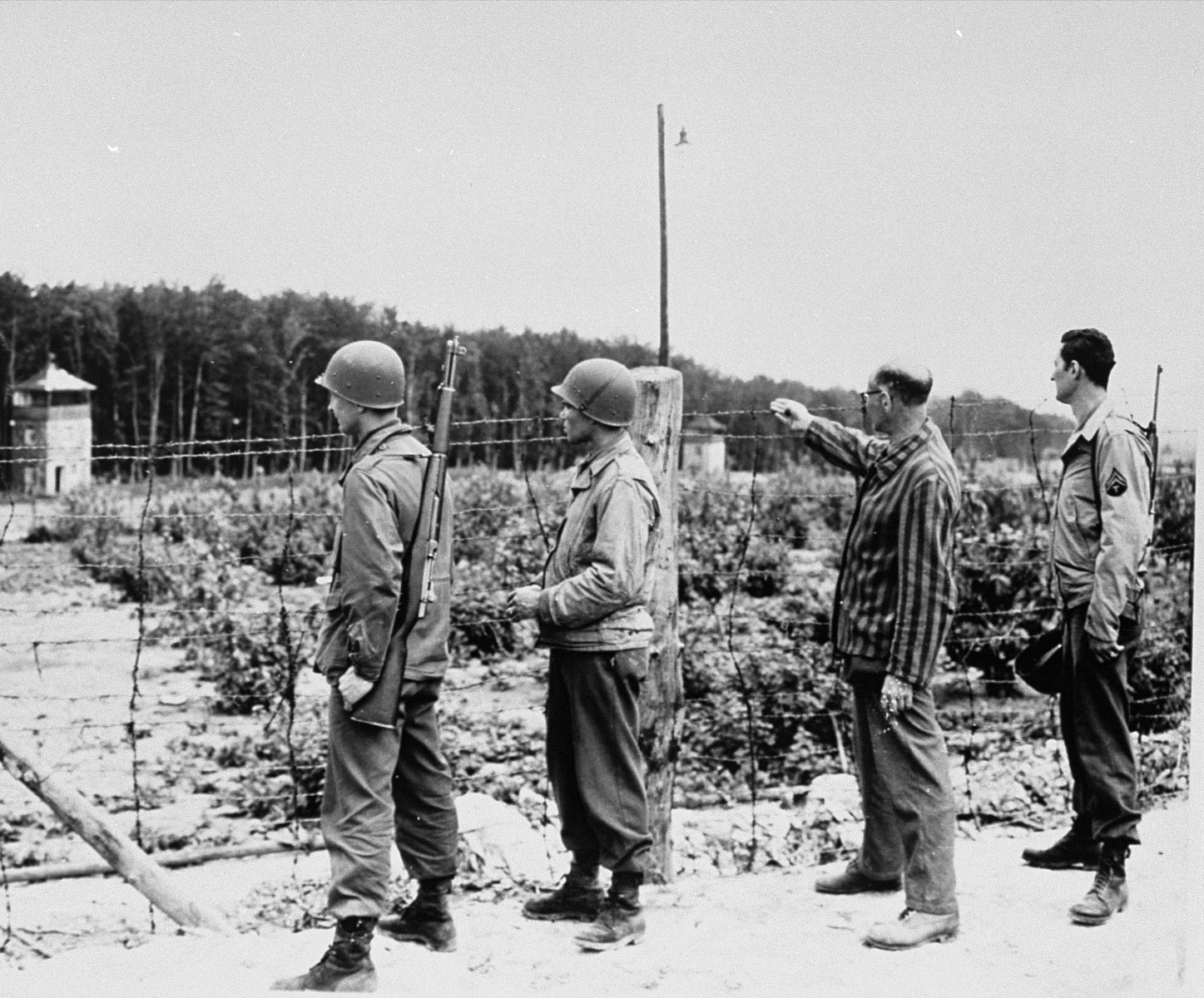 A survivor shows American troops of the 46th Armored Division, U.S. Ninth Army, the watch towers and the electrically charged barbed wire fence in the Buchenwald concentration camp.