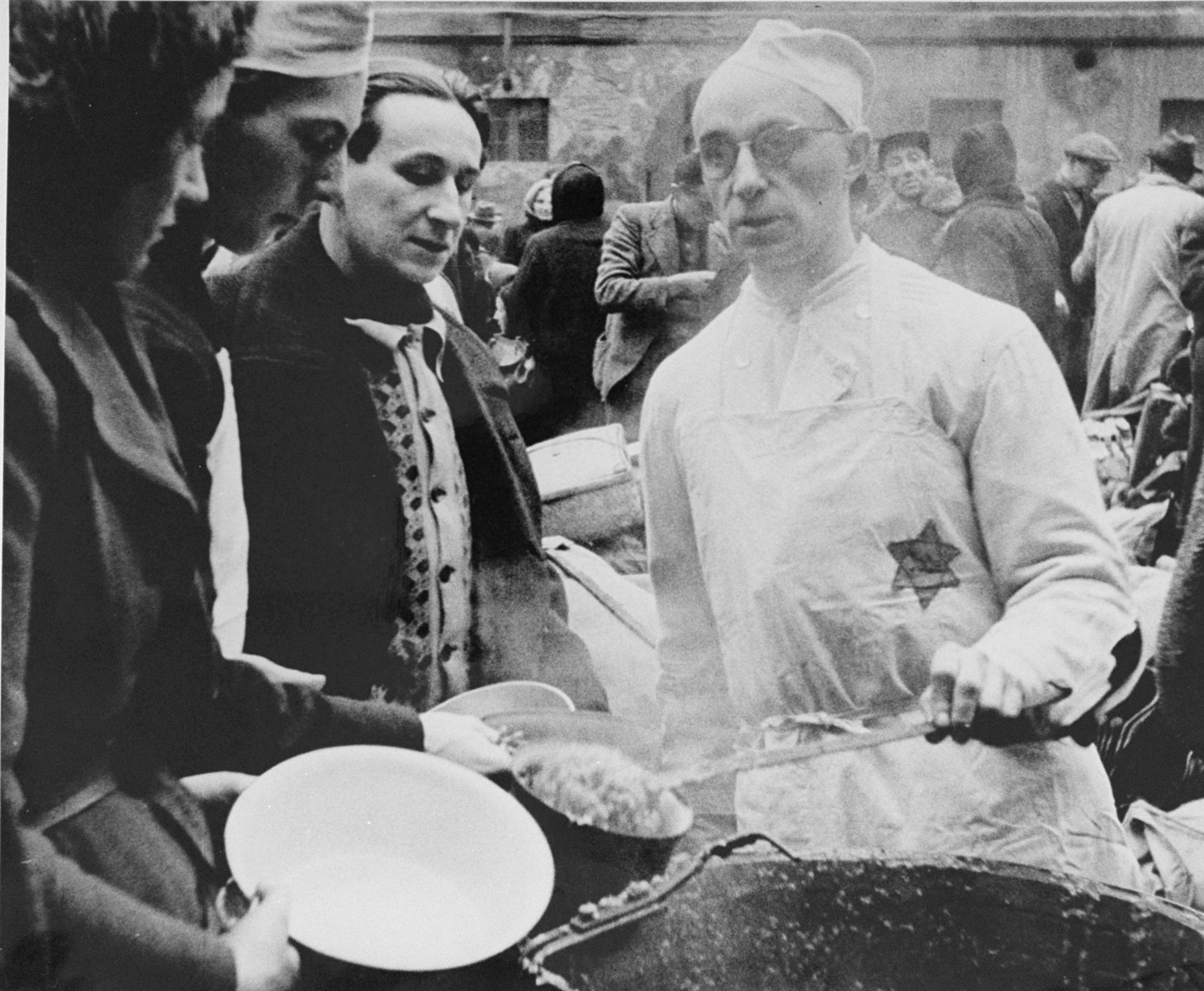 A prisoner wearing a cook's uniform doles out food in the ghetto courtyard to prisoners who have just arrived in Theresienstadt with a transport of Dutch Jews.  [oversized photo] 

The man serving soup is possibly Karel Arnstein.