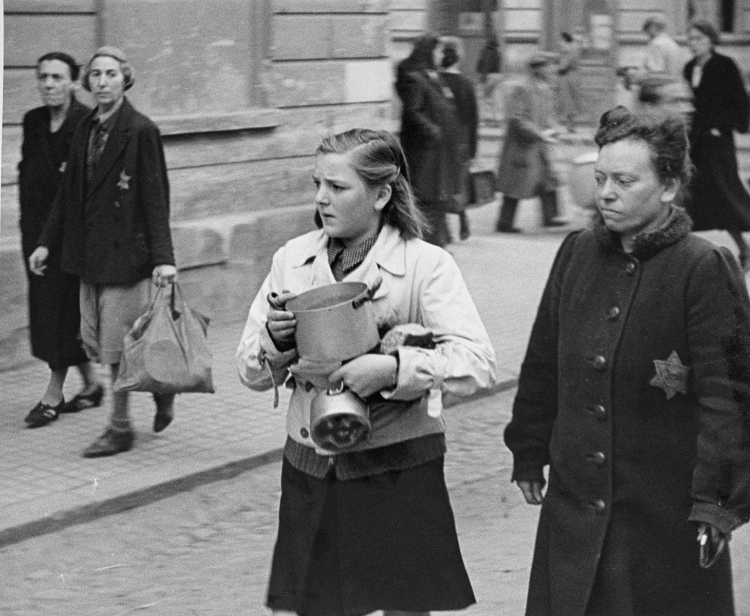 A teenage girl carries two small pots along a street in Theresienstadt.  Some of the Jews pictured appear to be new arrivals. [oversized photo]