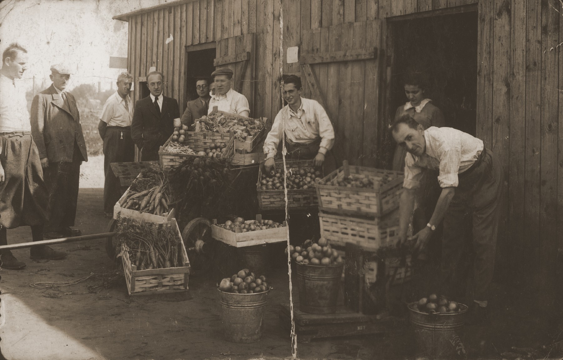Jewish workers put out their produce in boxes and pails in front of a wooden shed.
