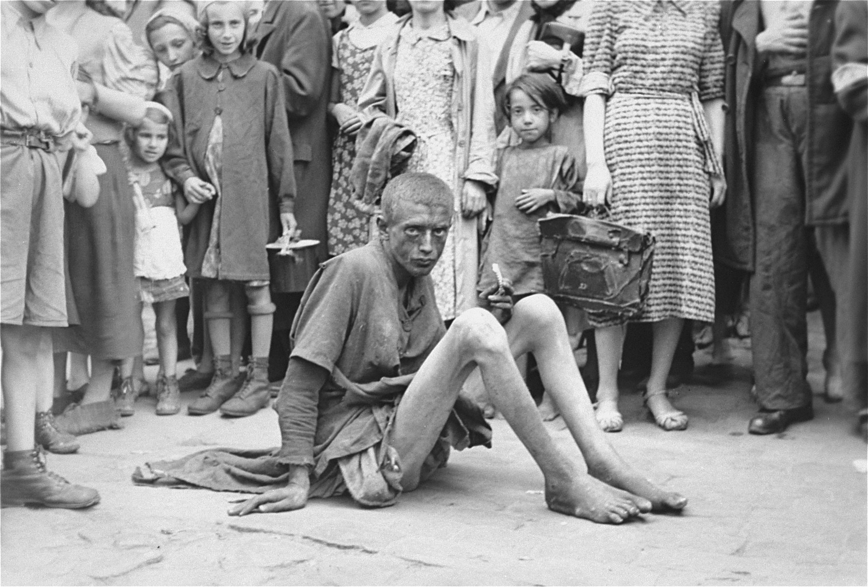 A destitute youth sits on the pavement in the Warsaw ghetto surrounded by other Jews.