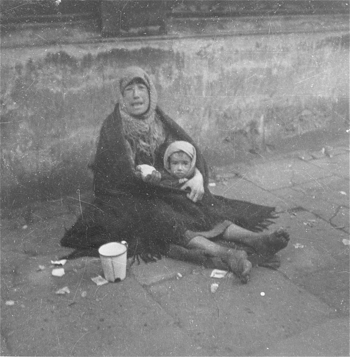 A destitute mother holding her child begs on a street in the Warsaw ghetto.  

Joest's original caption reads: "With a gesture of her bandaged hand this woman wanted to show me that her child had nothing to eat.  She wore a headscarf wrapped in a blanket but had no shoes."