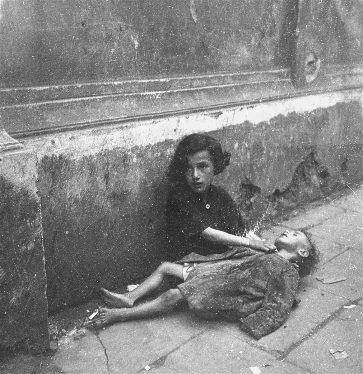 A destitute little girl comforts her little sister, who lies unconscious in her lap.  

Joest's original caption reads: "These were clearly sisters.  If the younger one was dead, I could not say.  She did not move."