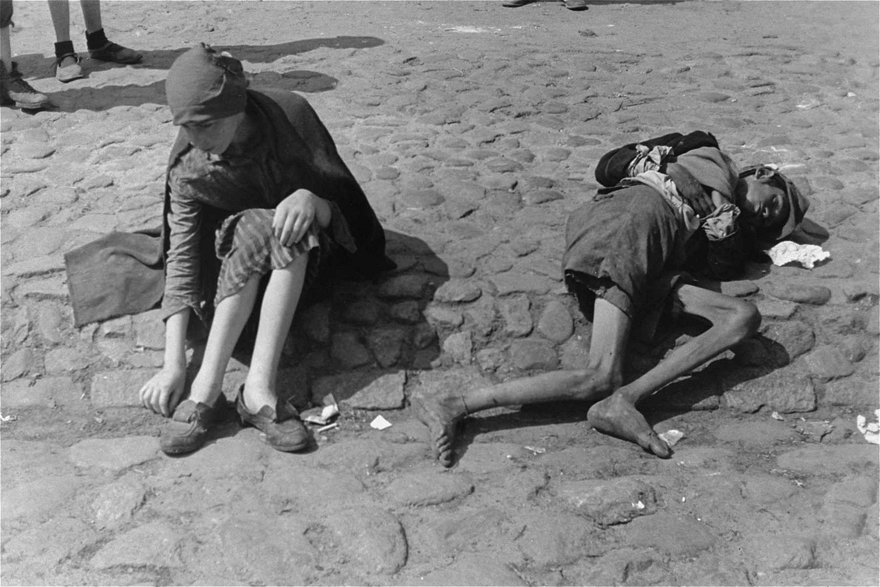 Two destitute children sit on the cobblestone pavement in a square in the Warsaw ghetto.