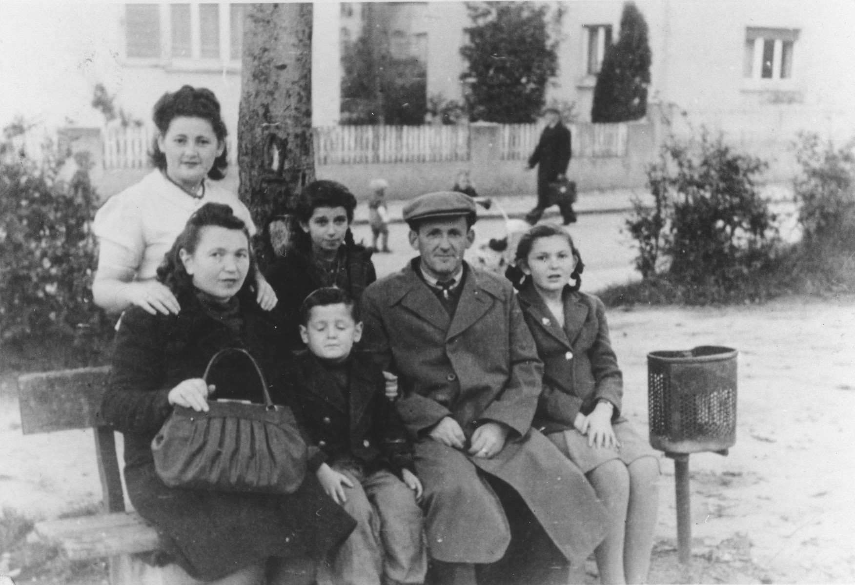 The Rajs family poses on an outdoor bench in the Ulm displaced persons camp.

Standing left to right are Chuma Rajs and Yanka Galler.  Seated left to right are Chaja, Srulek, Hershel and Lila Rajs.