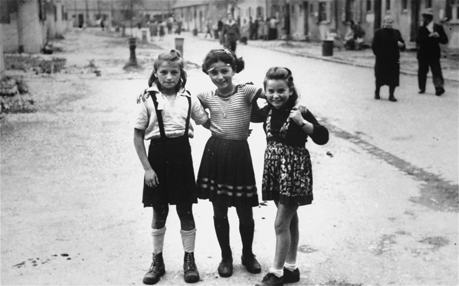 Three young girls pose together on a street of the  Foehrenwald displaced persons camp.  

Pictured from left to right are Pesa Balter, Chaia Libstug and Pela Roset.