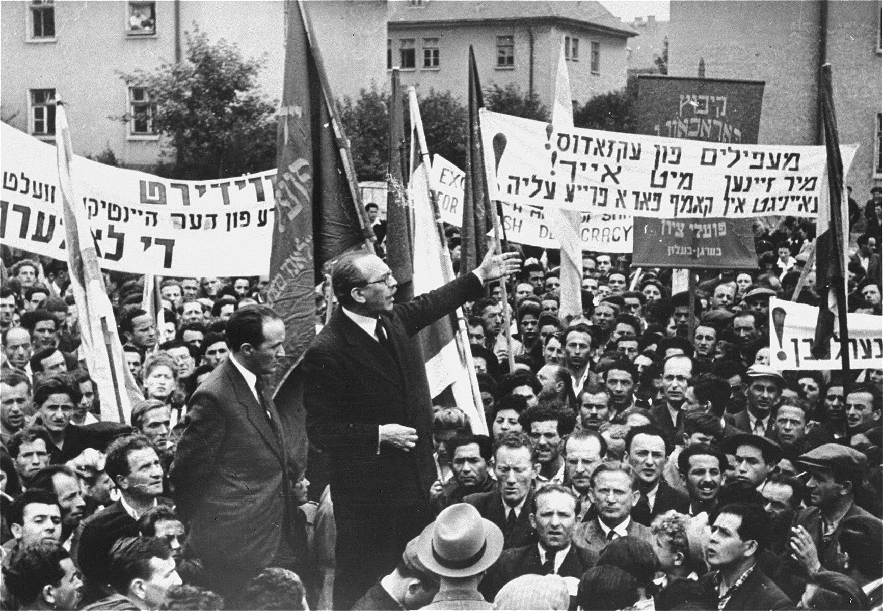 Marc Jarblum addresses a crowd of DPs at a demonstration protesting the forced return of the Exodus 1947 passengers to Europe.   

The Yiddish text of the banner on the right reads:  "Illegal immigrants from the Exodus we are with you!  United in the struggle for unrestricted immigration!"  
Josef Rosensaft stands next to the speaker.  Also pictured are Berel Laufer, Norbert Wollheim, and Max Silbernik.

Josef Rosensaft (1911-1975) was the son of Menachem Mendel and Devora (Szpiro) Rosensaft.  He was born and raised in Bedzin, Poland, where his father, a trained accountant, worked in the family's scrap metal business.  Josef had four older siblings: Chava, Rachel, Mari-Mindl and Itzhak.  His mother died in a flu epidemic when he was eight or nine.  During his youth and early adulthood, Josef was active in the labor Zionist movement.  After he finished school he joined his father in the scrap metal business.  Josef remained in Bedzin during the first four years of the German occupation of Poland.  On June 22, 1943 he was put on a deportation transport to Auschwitz, but managed to escape by diving out of the train into the Vistula River.  Though wounded by German bullets during his escape, Josef made it back to Bedzin the following morning.  A few weeks later during the final liquidation of the ghetto, his father died in his arms of natural causes. Josef then fled to the nearby town of Zawierce to escape the deportations.  However, that same month he was again rounded-up and sent to Auschwitz-Birkenau.  Josef spent five months in the camp before being transferred to the Auschwitz sub-camp of Lagisza Cmentarna in January 1944.  That winter he escaped from the camp and returned to Bedzin, where he was hidden by a Polish friend for six weeks.  He was recaptured in April and sent back to Auschwitz, where he was imprisoned in the Block 11 punishment barracks for seven months.  In November Josef was transferred to the Buchenwald sub-camp of Langensalza, and from there to Dora-Mittelbau early in 1945.  He was moved one last time to Bergen-Belsen in early April, where he was liberated by the British on April 15.  Within days of the liberation, Josef was chosen by his fellow survivors to become chairman of the Bergen-Belsen camp committee.  Later, after the convening of the First Congress of Liberated Jews in the British Zone in September 1945, he was elected chairman of the Central Jewish Committee for the British Zone of Germany.  He headed both the camp committee and the central committee until the closing of the Bergen-Belsen displaced persons camp in the summer of 1950.  During his years of leadership in the DP community Josef repeatedly stood up to the British in defense of the needs and political sensitivities of Jewish survivors.  He demanded that the British formally recognize Jews as a separate category of displaced persons.  He halted the transfer of Belsen DPs to two inferior camps established near the Dutch border.  He thwarted the British attempt to change the name of the Belsen DP camp to Hohne, which would have diminished the moral and political power of the DPs living there that derived from their link to the infamous concentration camp.  Finally, he repeatedly spoke out against the anti-Zionist policies of the British government and actively aided the illegal Bricha and Aliyah Bet movements that strove to get Jews out of Eastern Europe and smuggle them into Palestine.  On August 18, 1946 Josef Rosensaft married Hadassah Bimko (1912-1997), a fellow Bergen-Belsen survivor from Sosnowiec, Poland, who at the time was head of the health department of the Central Jewish Committee in the British Zone.  Hadassah was the daughter of Hersh Leib and Hendla Bimko, Gerer Hasidim who worked in the jewelry manufacturing business.  She had a younger brother, Benjamin and a younger sister, Roszka.  In the summer of 1943 Hadassah was deported to Auschwitz-Birkenau, where she spent fifteen months before being transferred to Bergen-Belsen in November 1944.  While she survived, most of her relatives, including her husband, son, parents and sister, perished in Auschwitz.  A dentist by training who had also studied medicine, Hadassah was recruited immediately after the liberation to organize a group of Jewish doctors and nurses in the camp to help British medical personnel treat the thousands of Belsen survivors suffering from disease and malnutrition.  Hadassah also served as one of the principal prosecution witnesses at the Bergen-Belsen Trial in Lueneburg (September-November 1945).  A year-and-a-half after their marriage, Hadassah gave birth to their son Menachem on May 1, 1948.  With the closing of the Belsen DP camp, the Rosensafts moved to Switzerland, where they spent eight years in the town of Montreux.  The family then immigrated to the United States, where Josef remained active in a variety of Jewish organizations.  He served as president of the World Federation of Bergen-Belsen Survivors until his death.