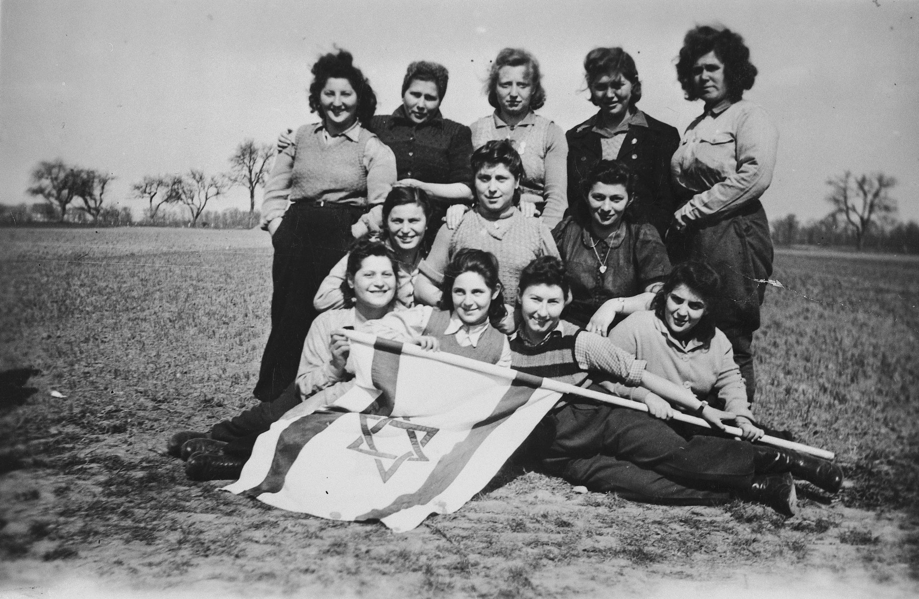 A group of young Jewish DPs who are seeking to immigrate to Palestine illegally, pose outside in a field with a Zionist flag.

Pictured in the front row from left to right are Kicia, Renia Klugman, Sabina Szeps, Sara Klugman; middle row: Ester Goldschmidt, Mina Kokodinska, Hanka Wiernik Schlesinger; top row: Goldi Silberstein, unknown, Dora, unknown, Bracha.