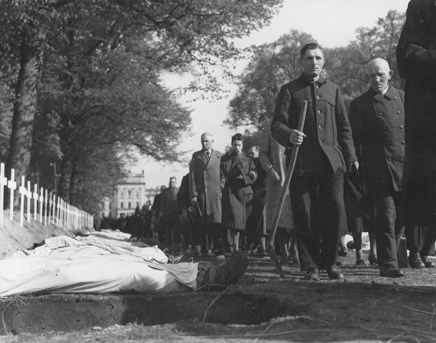 Upon the orders of the U.S. Army, the population of Ludwigslust files past the bodies of prisoners killed in the Woebbelin concentration camp, which have been laid out for burial in individual graves on the palace grounds of the Archduke of Mecklenburg.  The men carry shovels to bury the victims.