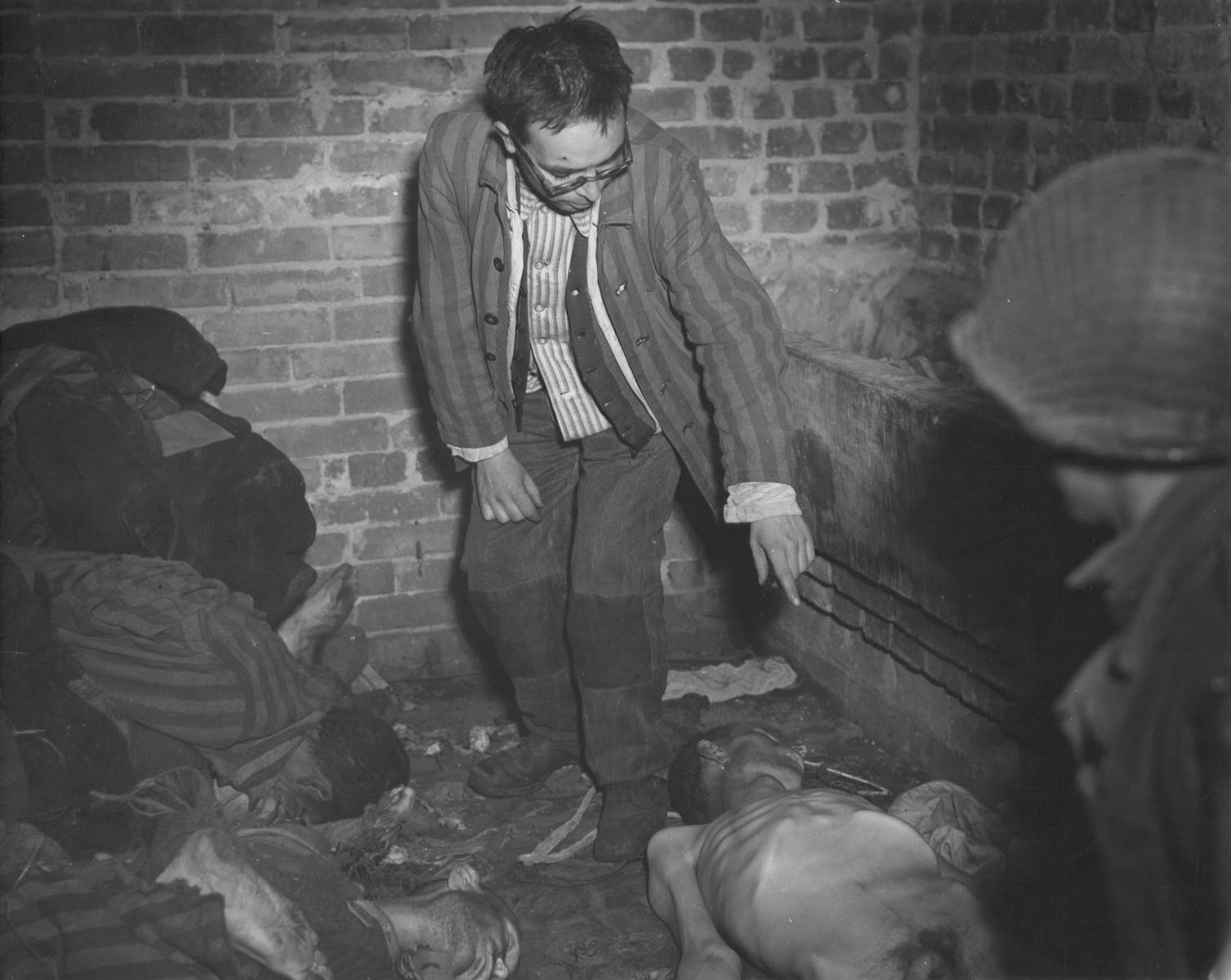 David Rousset, a liberated prisoner who is acting as a guide for an American soldier, points out the emaciated corpse of a prisoner lying on the floor of a brick structure in the newly liberated Woebbelin concentration camp.