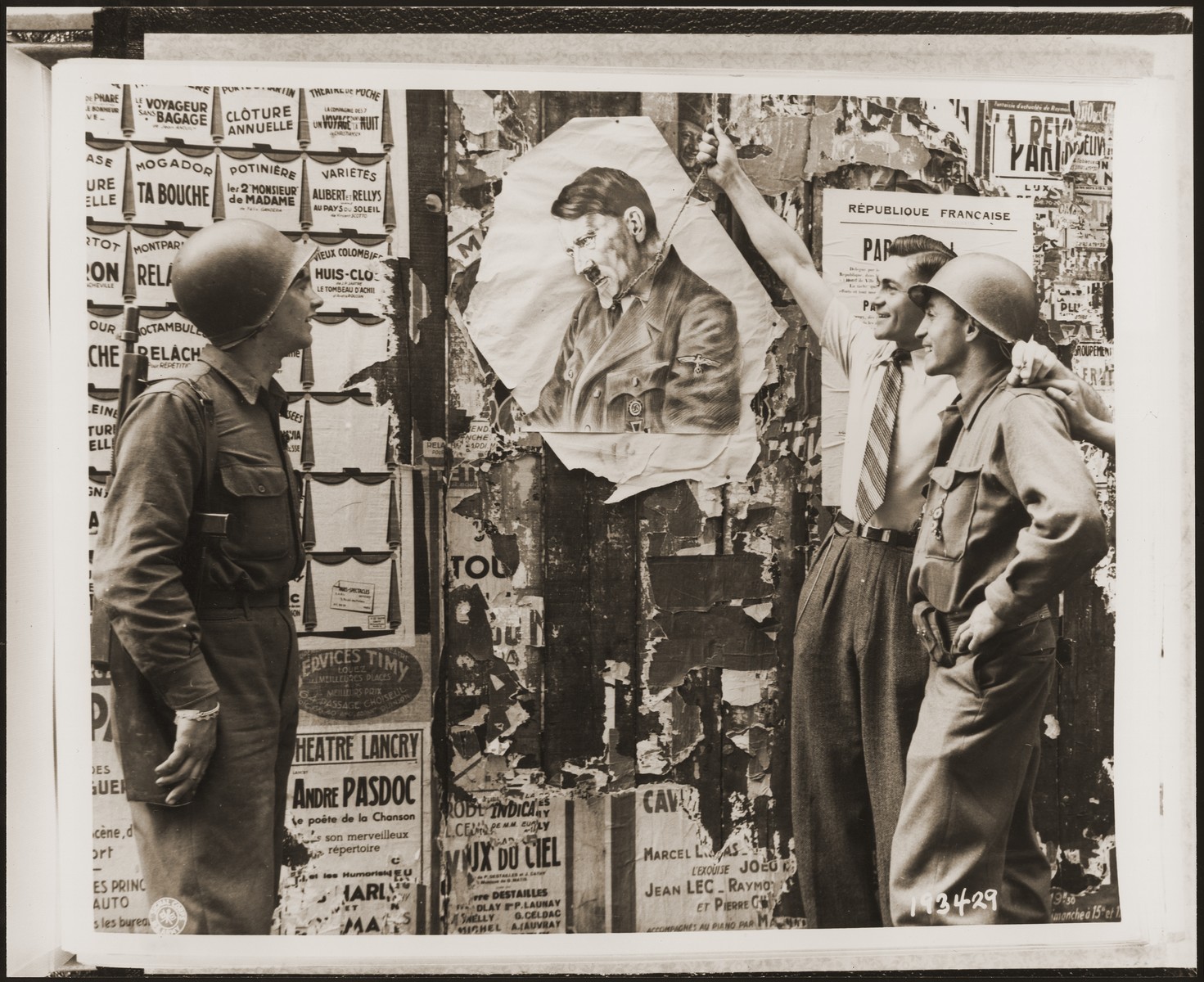 Two American soldiers watch as a Frenchman "hangs" in a portrait of Hitler in effigy.