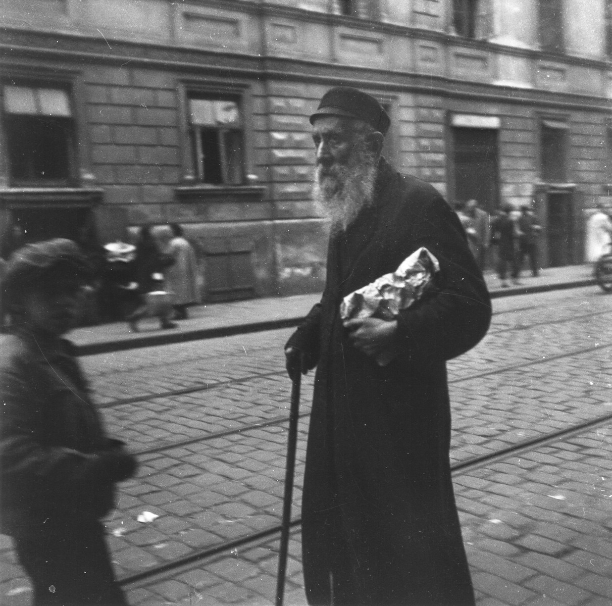 An elderly Jew holding a package and a cane walks along a commercial street in the Warsaw ghetto.  

Joest's original caption reads: "It struck me like an image out of the Old Testament--this old man with a small bundle in his arm, walking towards certain death."