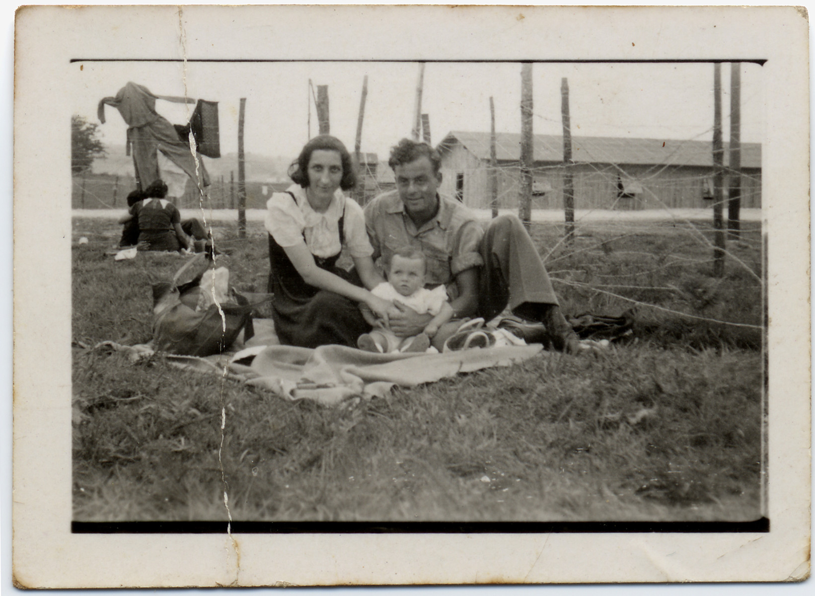 A couple with their young child sits on the grass near the barbed wire fence of the Gurs internment camp. 

Pictured are Tonia and Sioma Lechtman with their baby Vera.