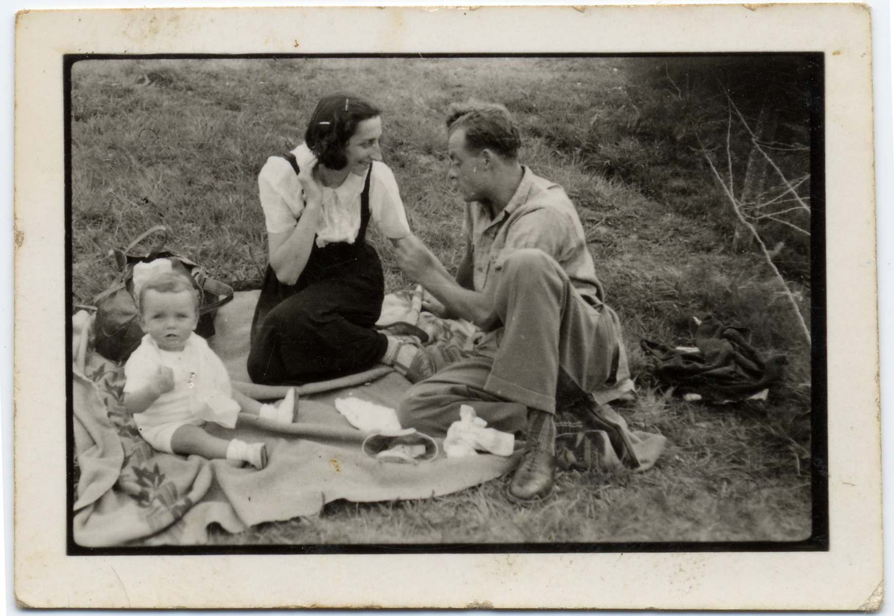 A couple with their young child sits on the grass on an open field in the Gurs internment camp. 

Pictured are Tonia and Sioma Lechtman with their baby Vera.