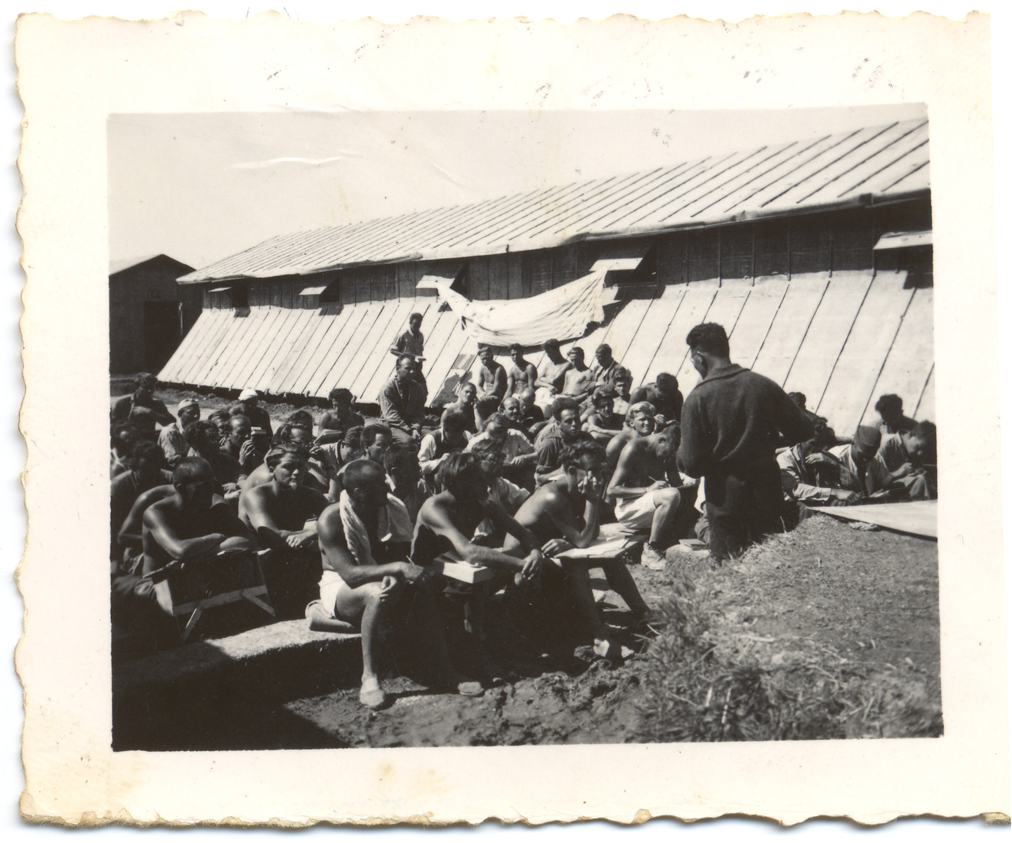 Prisoners [possibly members of the International Brigade who fought in the Spanish Civil war] listen to a speaker in the Gurs internment camp. 

The donor's father might be the man sitting on the far right and taking notes.