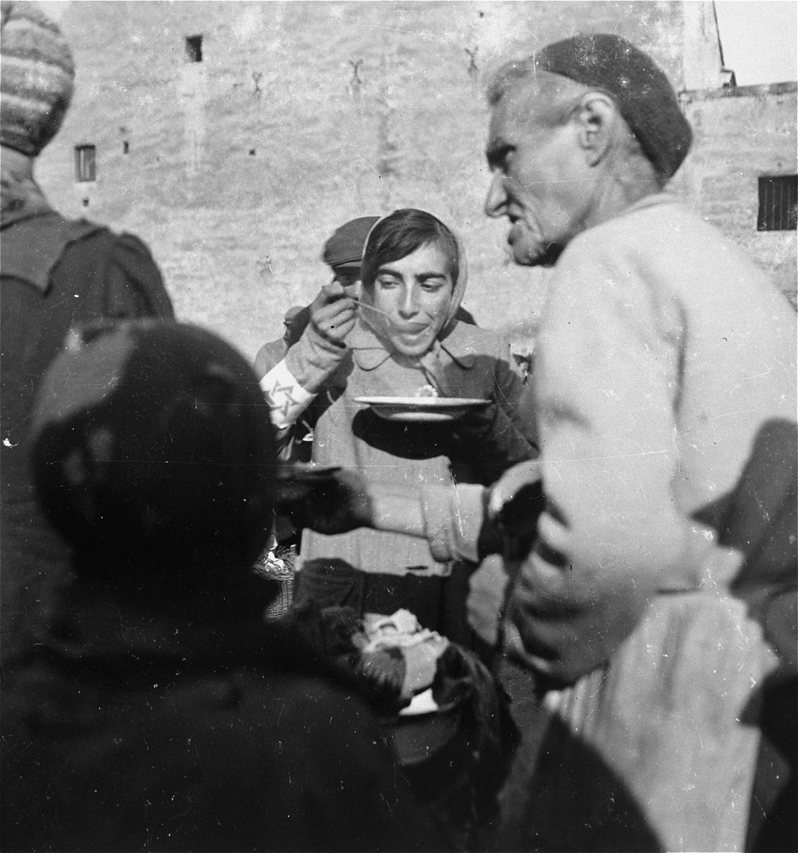 A woman in the Warsaw ghetto eats soup she received from a public kitchen.  

Joest's original caption reads: "Food from a large tureen was given out to Jews who waited there in a long line.  The woman in the foreground stood in the line and held out her bowl.  The woman behind her ate her thin soup from a lead bowl."