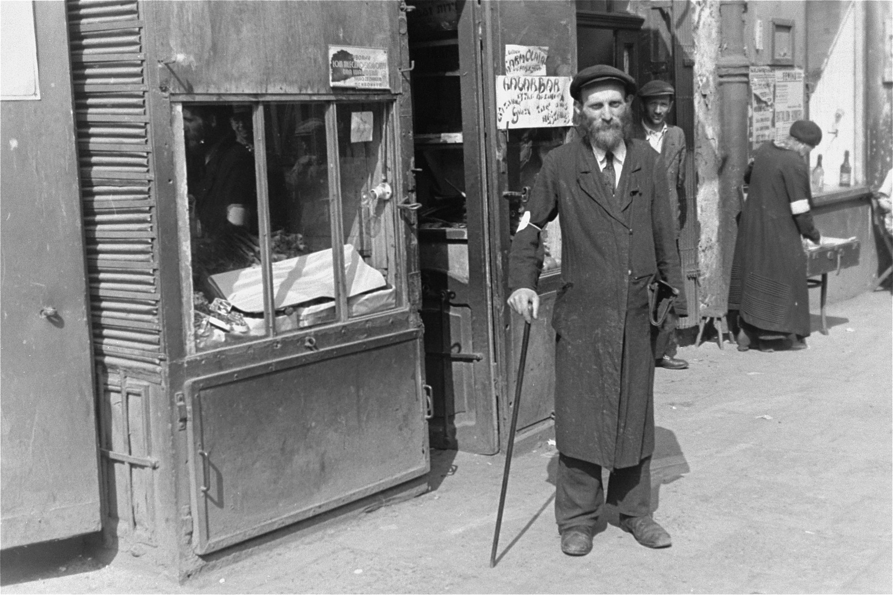 An Jewish man walks along a street in the Warsaw ghetto with a cane.