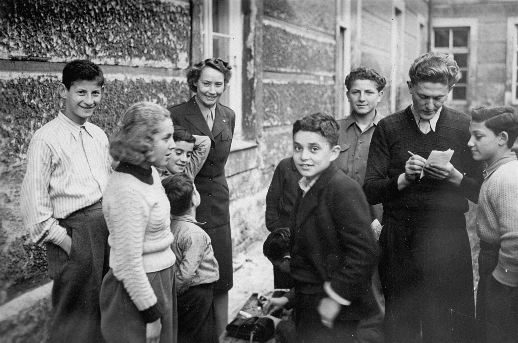 UNRRA relief worker Marion E Hutton talk to some of the children in front of the Kloster Indersdorf DP children's center.

Wladislaus Fischer is on the far left.  Nina Krieger is second from the left.  In the middle is Steve Israeler.  Also pictured partially obscured are the two Weinstock brothers.  Mioses Sztajnkeler is in the center right facing the camera and Dezider Kahan is second from the right.