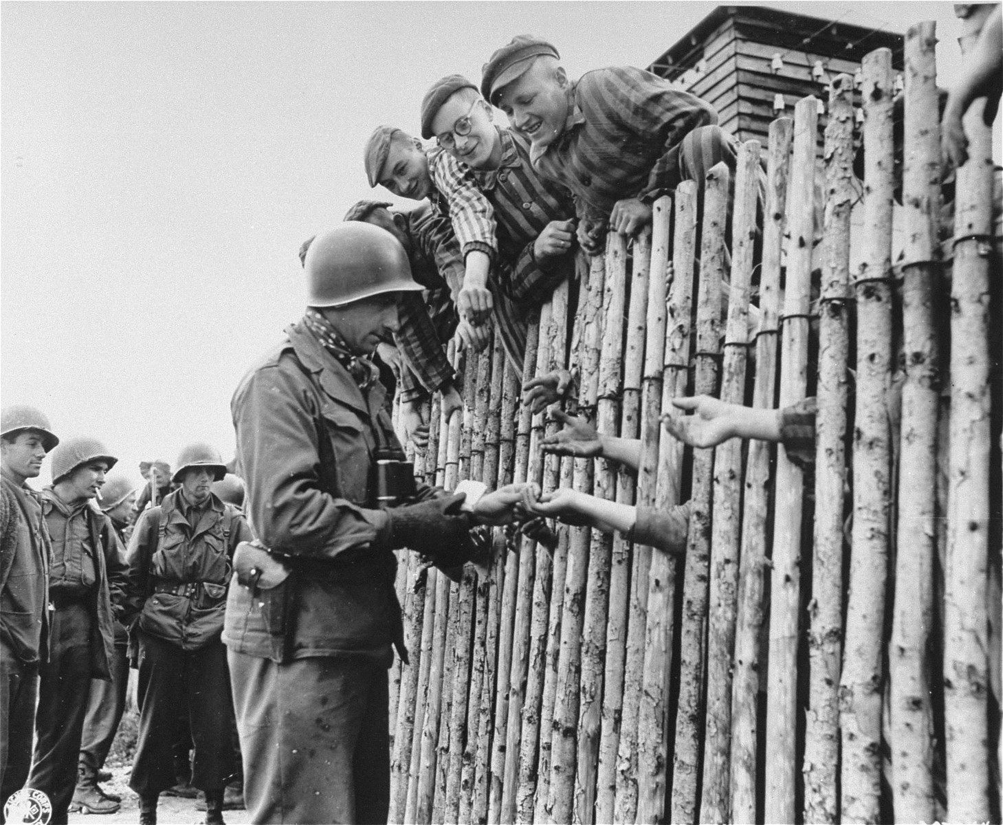U.S. Army Corporal Larry Matinsk puts cigarettes into the extended hands of newly liberated prisoners behind a stockade in the Allach concentration camp.

Cpl Larry Mutinsk served in  Company A, 1st Battalion, 157th Infantry Regiment, 45th Infantry Divison.  Also pictured are U.S. Army soldiers Arthur Toratti and George Babel (second and third from the left).

Original caption reads, "Cpl Larry Mutinsk, Philadelphia, PA., hands out his last pack of cigarettes to the eager reaching hands of the prisoners within the wire stockade of the prison camp at Dachau."