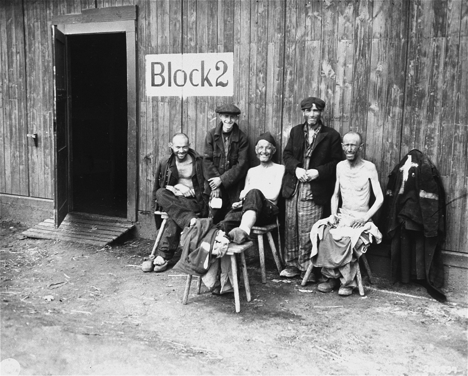 Five Jewish survivors pose for a U.S. Signal Corps photographer in front of Block 2 in the Hannover-Ahlem concentration camp, a sub-camp of Neuengamme.  

The man second from left is Sam Gottesman.

The original caption reads "Liberated, a concentration camp near Hanover, Germany, these five Jews smile happily despite their starved and ill condition."

The original Signal Corps caption reads, 
"PNA                                                          EA61998

U.S. TROOPS LIBERATE PRISONERS OF NAZIS
Five sick and undernourished Jews are shown before one of the barracks of the Nazi Hanover-Harlen concentration camp near Hanover, Germany, following capture of the city by Ninth U.S. Army troops April 10, 1945.  Hanover is 155 miles west of Berlin.  U.S. Signal Corps Photo ETO-HQ-45-31748.
SERVICED BY LONDON OWI TO LIST B-1
CERTIFIED AS PASSED BY SHAEF CENSOR