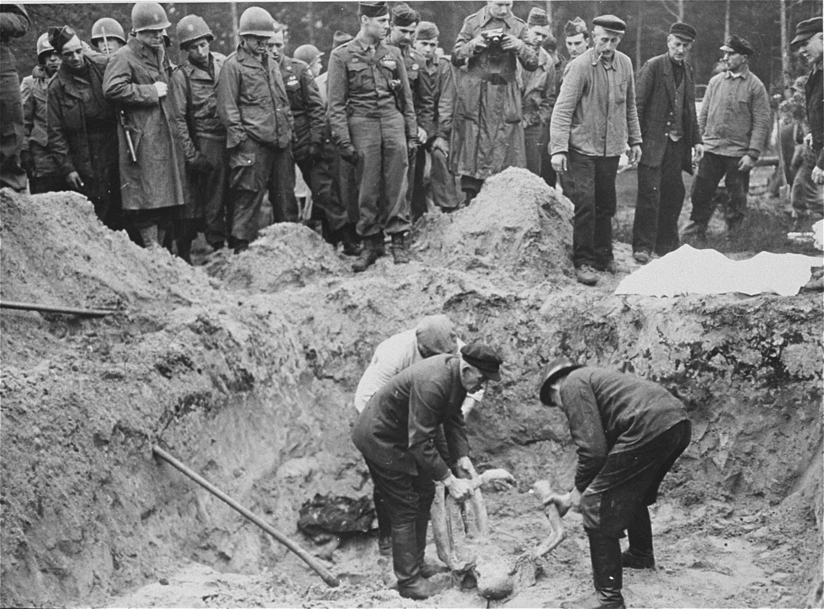 American troops with the 82nd Airborne Division look on as German exhume corpses from a mass grave. 

The original Signal Corps caption "NEW NAZI HORROR CAMP DISCOVERED.  One of the worst Nazi concentration camps uncovered by Allied troops was liberated at Wobbelin, Germany, a small town five miles north of Ludwigslust and 90 miles northwest of Berlin.  Soldiers of three Allied units -- the 82nd U.S. Airborne Division, the Eighth Infantry Division of the Ninth U.S. Army and airborne troops of the Second British Army -- entered the camp and found sick, starving inmates barely surviving under indescribable conditions of filth and squalor.  They found hundreds of dead prisoners in one of the buildings while outside, in a yard, hundreds more  were found hastily buried in huge pits.  One mass grave contained 300 emaciated, disfigured corpses.  The dead included Poles, Russians, Frenchmen, Belgians, Dutchmen and Germans, all of whom had been working as slave laborers for the Nazis.

It is estimated that at least 150 of the original 4,000 prisoners succumbed daily, mostly from starvation and savage treatment at the hands of Nazi SS troops who operated the camp.   Some of the bodies found were burned almost beyond recognition and systematic torture of the inmates was revealed by the  physical condition of most of the survivors.  Military Government officers immediately ordered leading citizens of nearby Ludwigslust and other towns to march through the camp and witness the atrocities committed by representatives of the German Government.  Most of the civilians disclaimed any knowledge of the camp's existence despite the fact that many of the prisoners worked in the area.

The local residents later were made to exhume the bodies from the mass graves at the camp and provide decent, respectable interment of all dead prisoners.  Two hundred were buried in the public square of Ludwigslust May 7, 1945, and an equal number were buried in the garden of the highest Nazi official of Hagenow.  Eighty more were laid to rest in the town of Schwerin.

BIPPA                                                          EA 66636

THIS PHOTO SHOWS:  German civilians of Ludwigslust and nearby communities are forced to exhume the bodies of the Nazi atrocity victims from a mass ... Wobbelin concentration camp.  These ... reburied the victims in the town ... gslust.  Troops of the 82nd U.S. ... witness and direct the proceedings ...
Photo ETO-..."