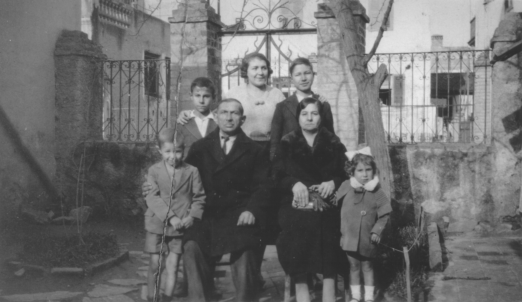Portrait of the Beraha family in the courtyard of their home in Salonika.  

Pictured are Eliahu and Sol Beraha (center) and their children: Yeshua (left), Alberto (back row, left), Miriam (back row, center), Baruch (back row, right) and a niece named Estrea (lower right).