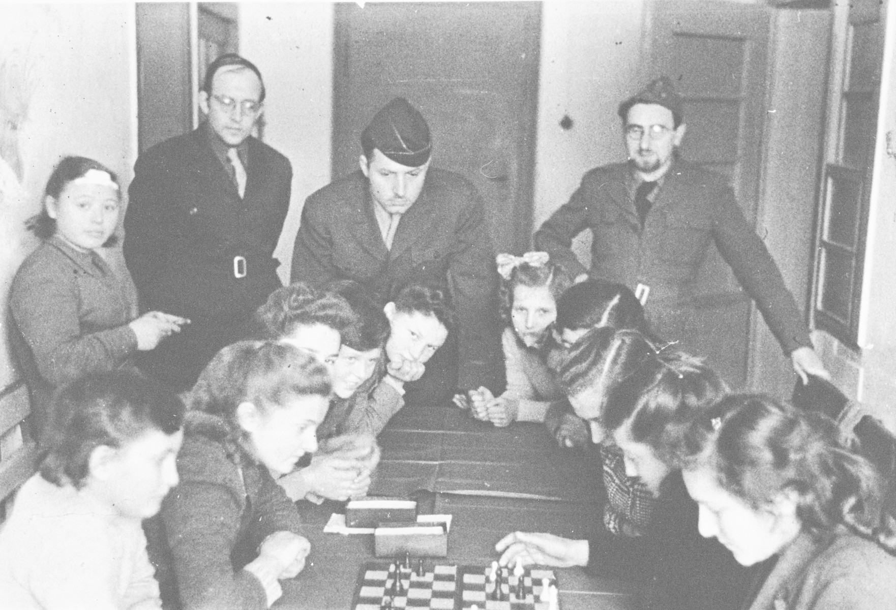 School girls play chess in the Poalei Agudat Yisrael children's home in Ulm run by the Vaad Hatzala while Vaad rabbis look on.

Rabbi Nathan Baruch is in the center and Rabbi Aviezer Burstin is standing on the right.
