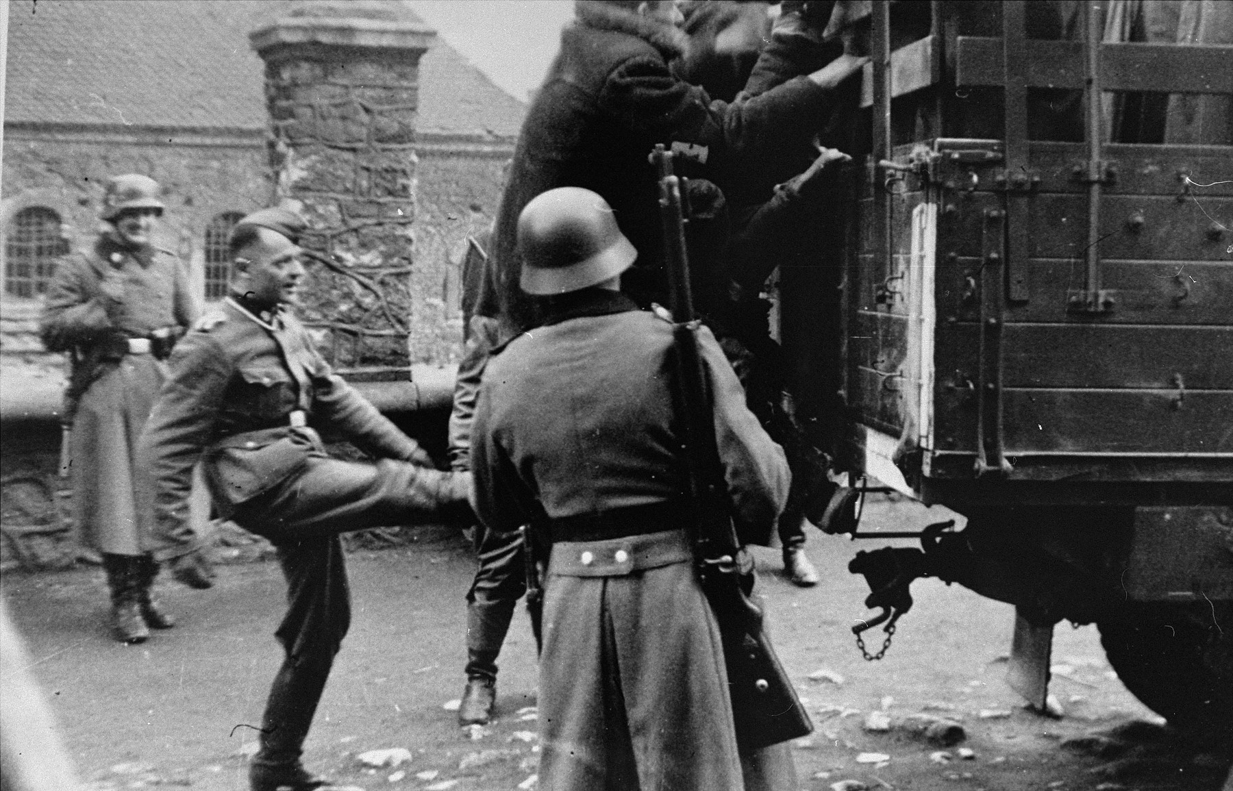 A member of the German police kicks a Jew who is climbing onto the back of a truck during a round-up for forced labor.  Two other Germans look on with derision.