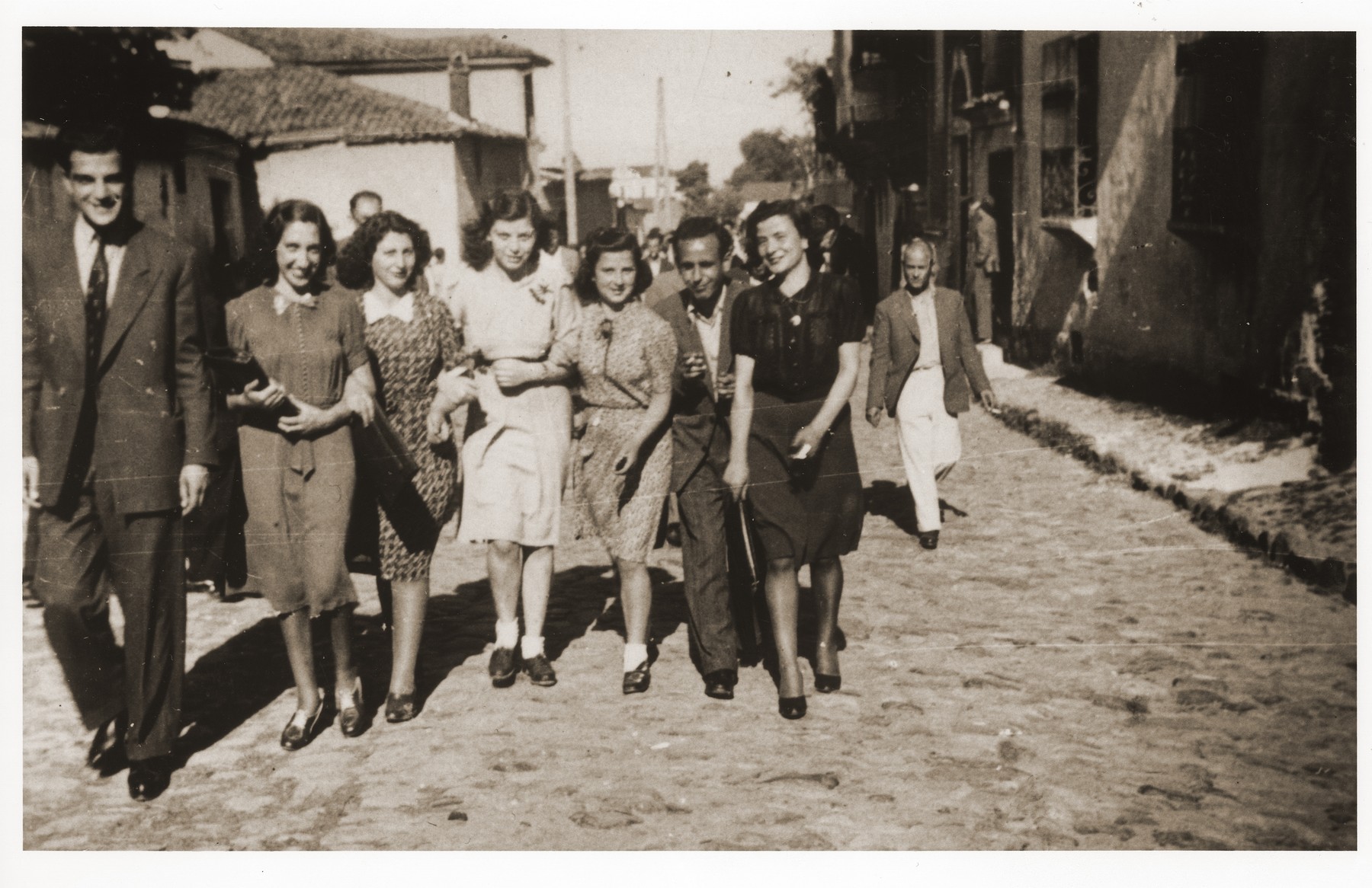 Family and friends walk down Tabaneh Street in the Jewish quarter of Monastir, just  after a wedding (synagogue pictured in the background on the left).

Among those pictured are (left to right) Leon Franco (a cousin of Rachel), Rachel (nee Nahmias) Kornberg, [first name unknown] Pardo, [first name unknown] Shami, Elinor [last name unknown], Moise [last name unknown], and [an unidentified woman].