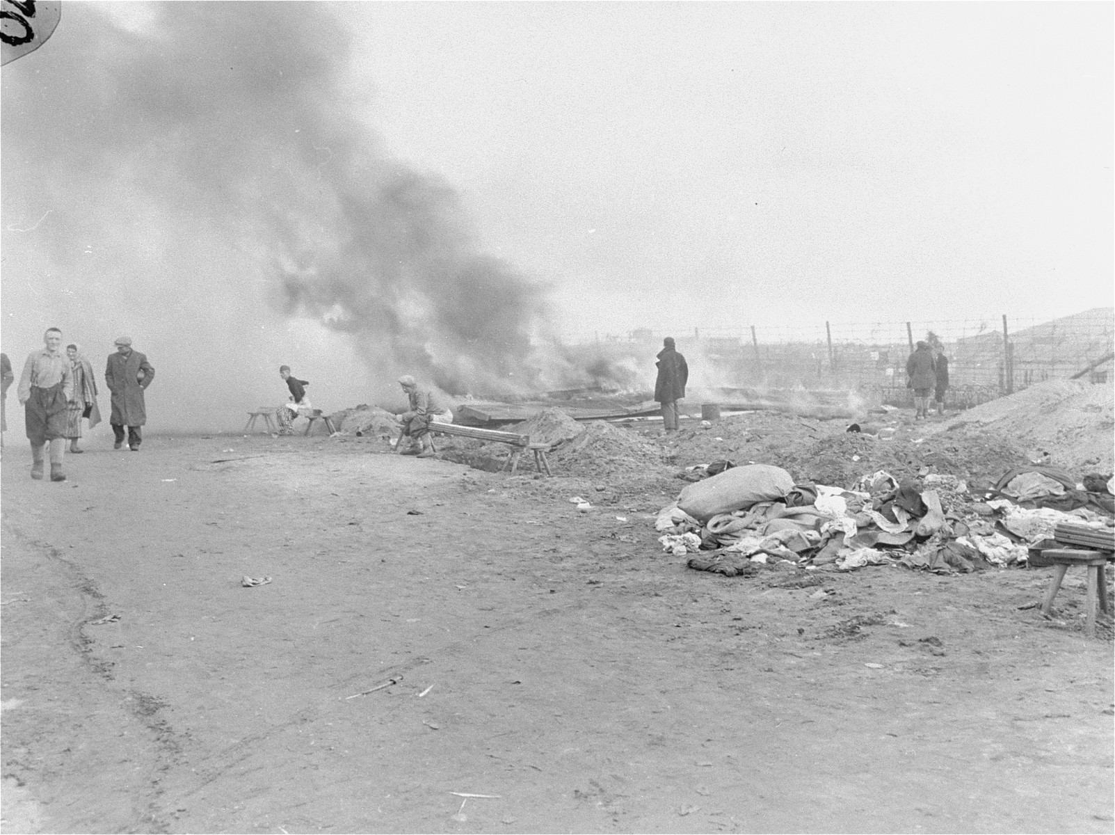Billows of smoke rise up to the sky from typhus-infested barracks in Bergen-Belsen that have been torched by the British military.  A group of survivors move about just in front of the smoke.