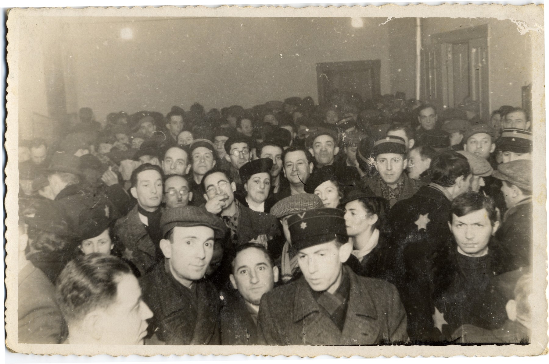 Jewish men and women wearing Stars of David, including several policeman, crowd together in a hall in the Lodz ghetto.