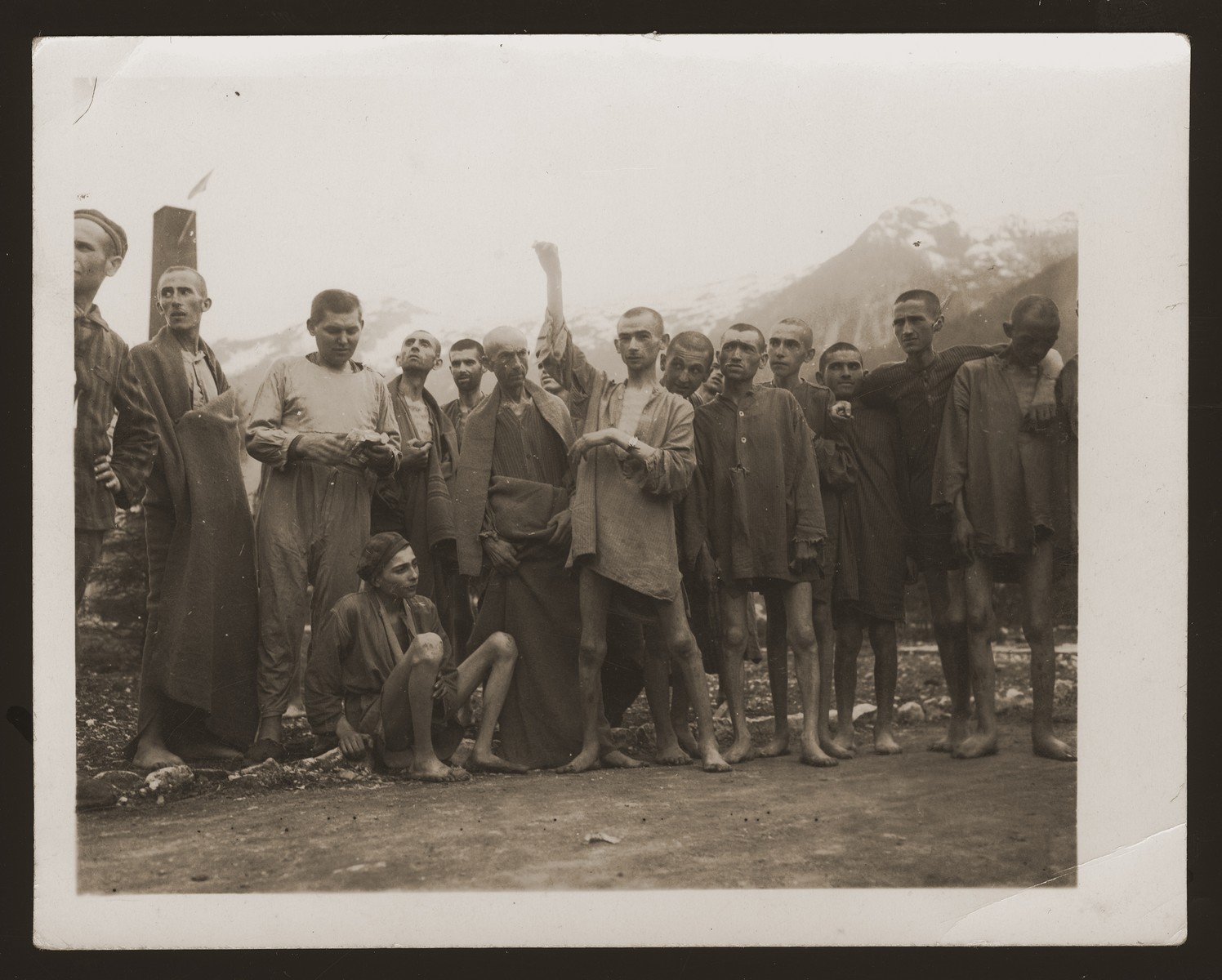 One day after their liberation, a group of former prisoners at the Ebensee concentration camp pose outside for US Army Signal Corps photographer Arnold Samuelson.

The former prisoners came out of the infirmary barracks in order to be photographed.  Visible behind them is the top of the crematorium chimney with a white flag on top.  Among those pictured is a 21-year-old Jewish survivor by the name of Joachim Friedner from Krakow, Poland (center with raised arm).