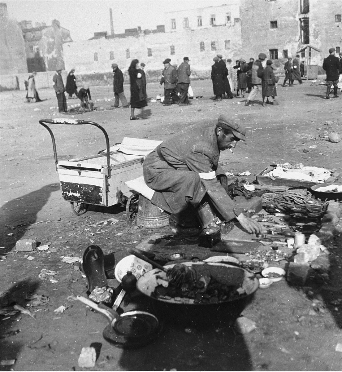 A man offers various items for sale on a lot in the Warsaw ghetto. 

Joest's original caption reads: "This man offered [for sale] nothing anyone could need: old pans, heavy, dented bowls, an old cauldron.  However, he had to try any way he could to make money to live."