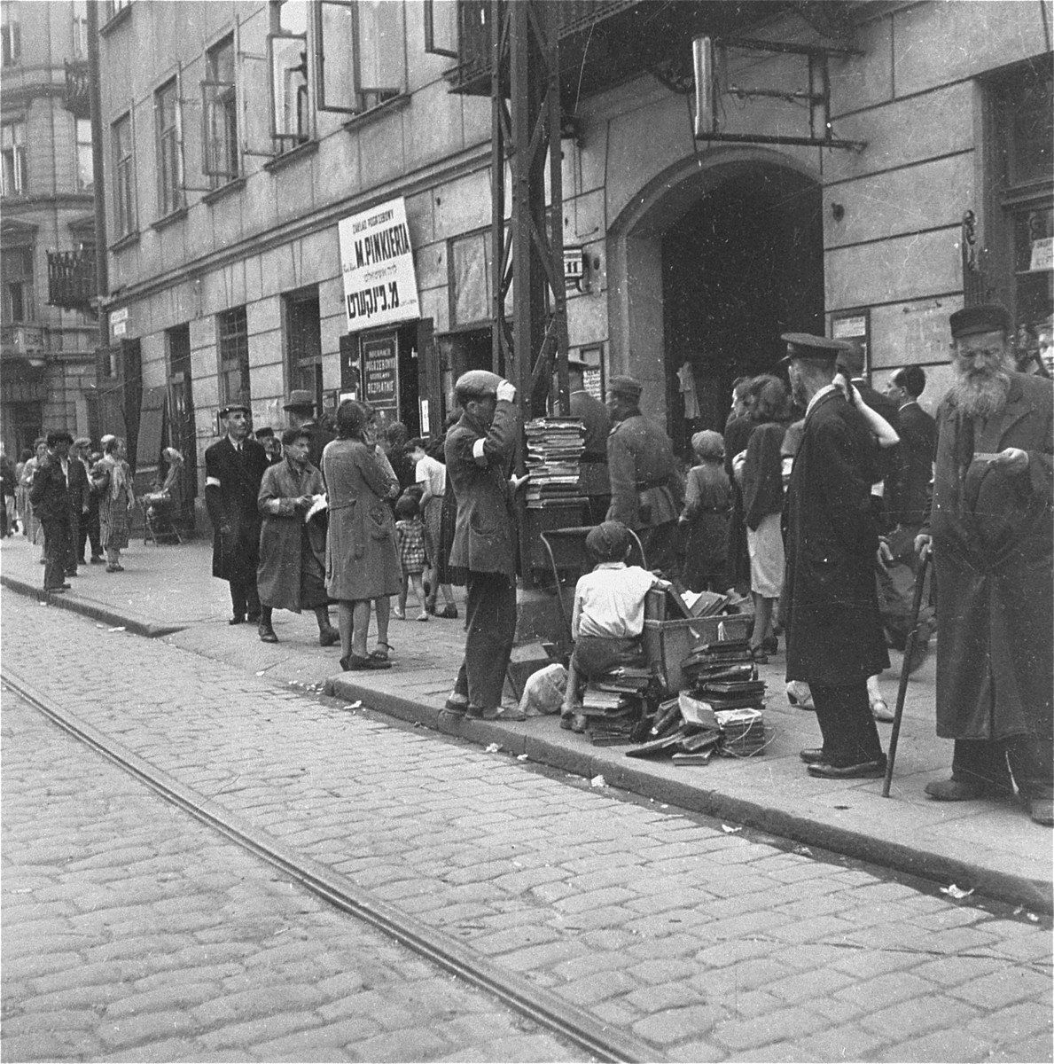 A vendor sells used books on Nowolipiki Street near the undertaker's business of Mordechai Pinkiert.  

Joest's original caption reads: "This was Nowolipiki Street, with the office of an undertaker.  I did not notice the name Mordechai Pinkiert at that time."