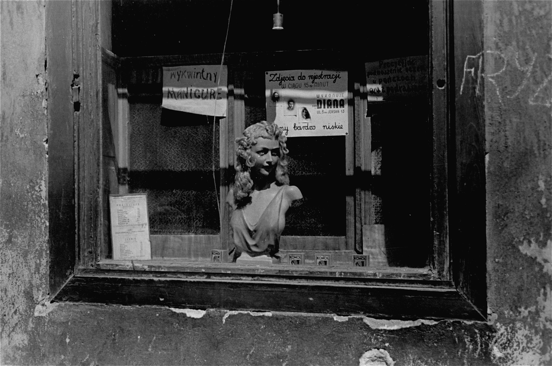 A shop window in the Warsaw ghetto advertising different services, including stocking repair, passport photos and manicures.  The chalk sign on the wall reads "barbershop."