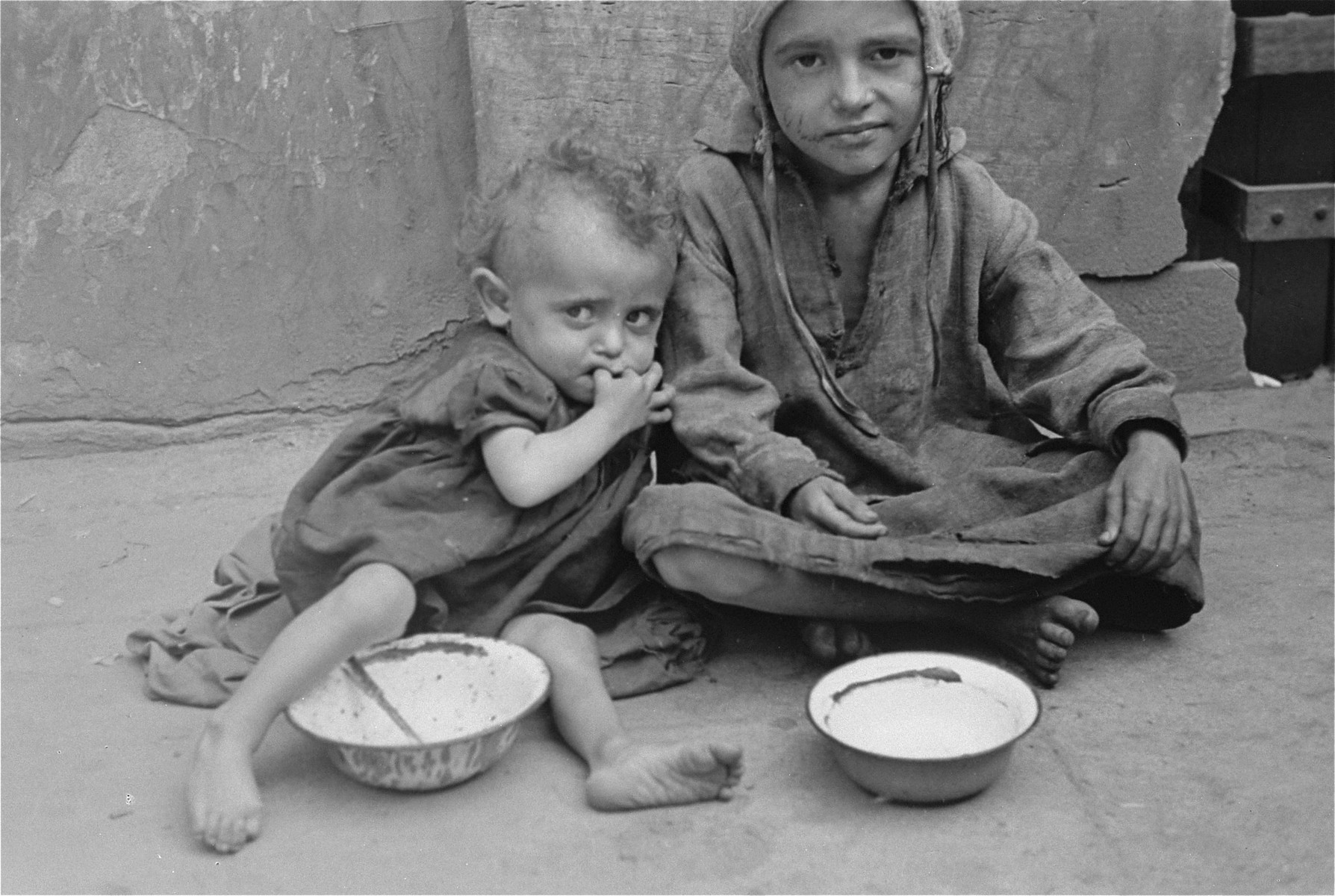 Two destitute children sit with empty bowls on a street in the Warsaw ghetto.