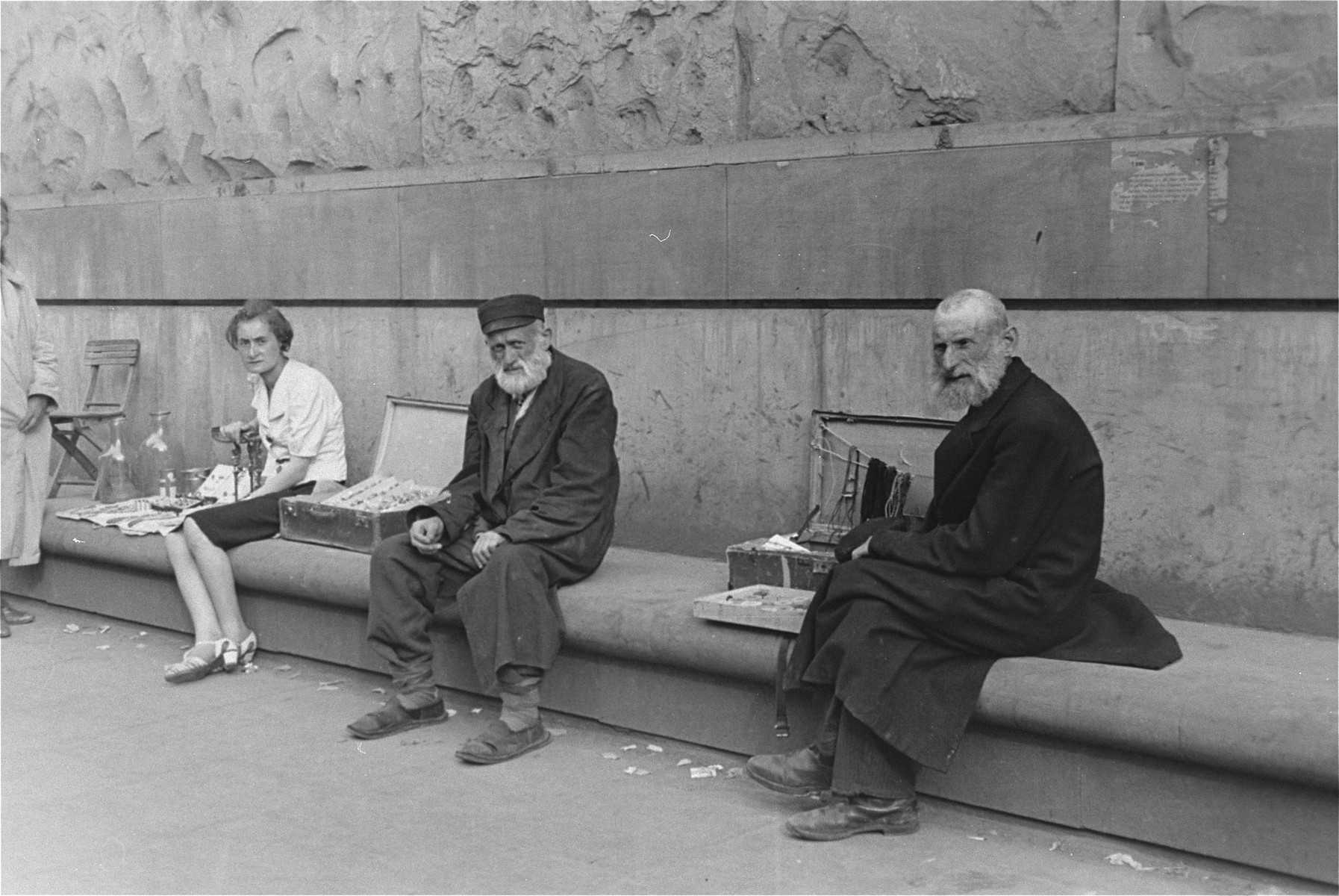 Three street vendors who are seated on a long bench, display their wares in the Warsaw ghetto.