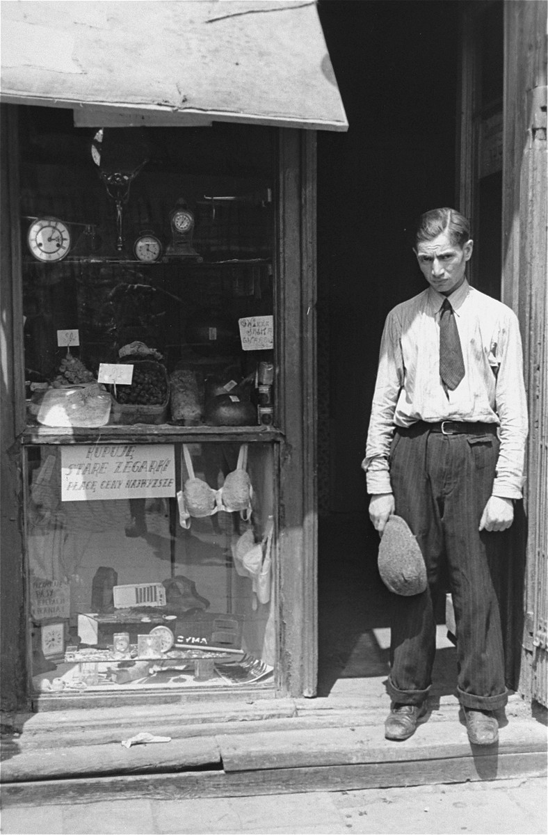 A young man stands in the doorway of a shop in the Warsaw ghetto.  He has taken off his hat in compliance with the German regulation that Jews remove their hats in the presence of German personnel.  The sign in the window reads: "Buying old watches, will pay top prices."