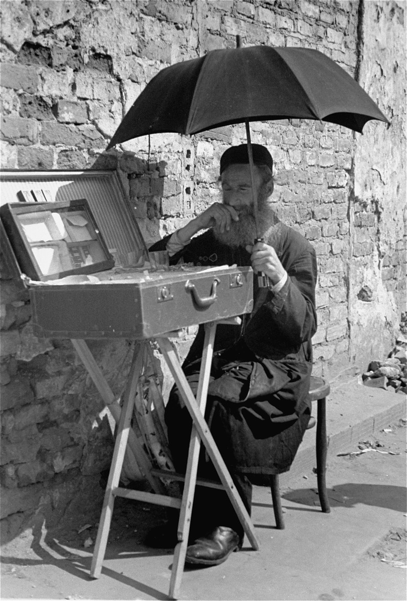 A Jewish vendor sitting on the street in the Warsaw ghetto shades himself with a large umbrella.