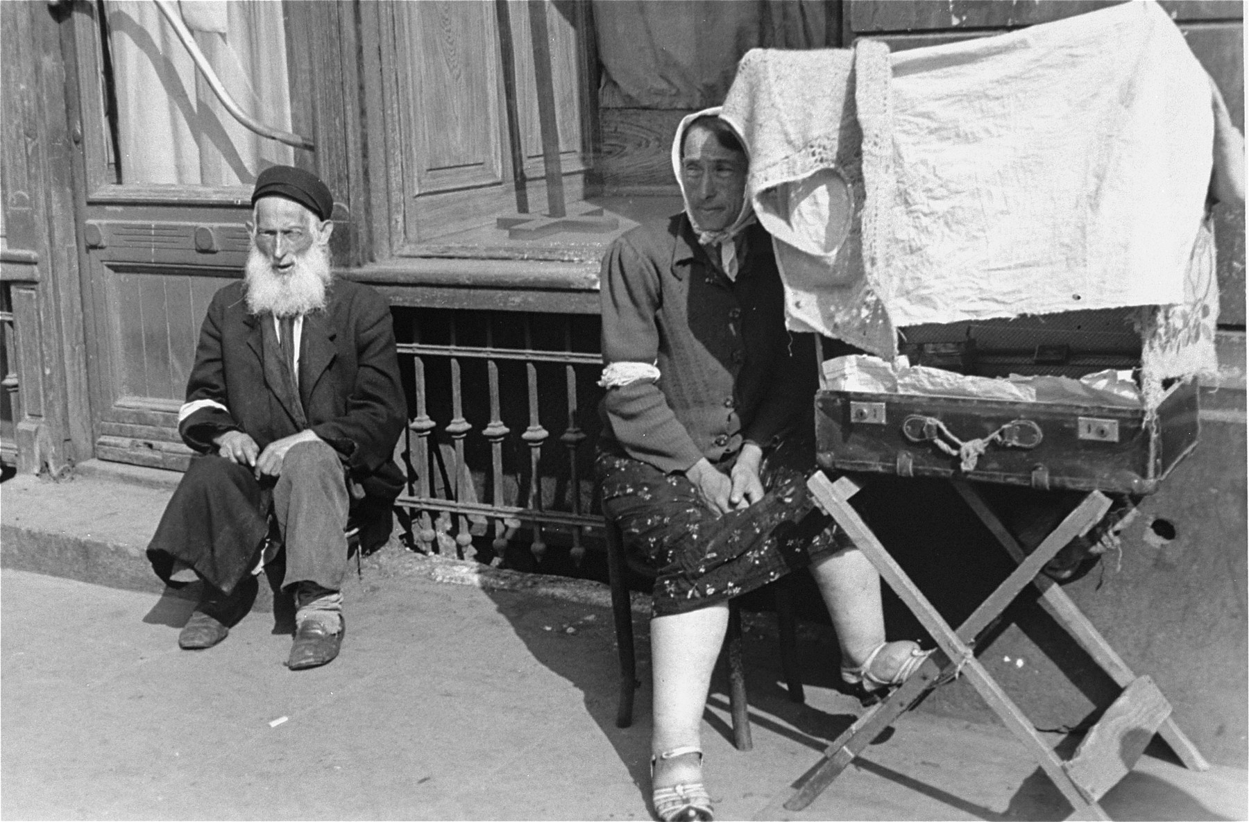 A female vendor sits on a street in the Warsaw ghetto next to an open suitcase containing her wares that has been covered with a dress to provide some shade.

An elderly, bearded man sits nearby.
