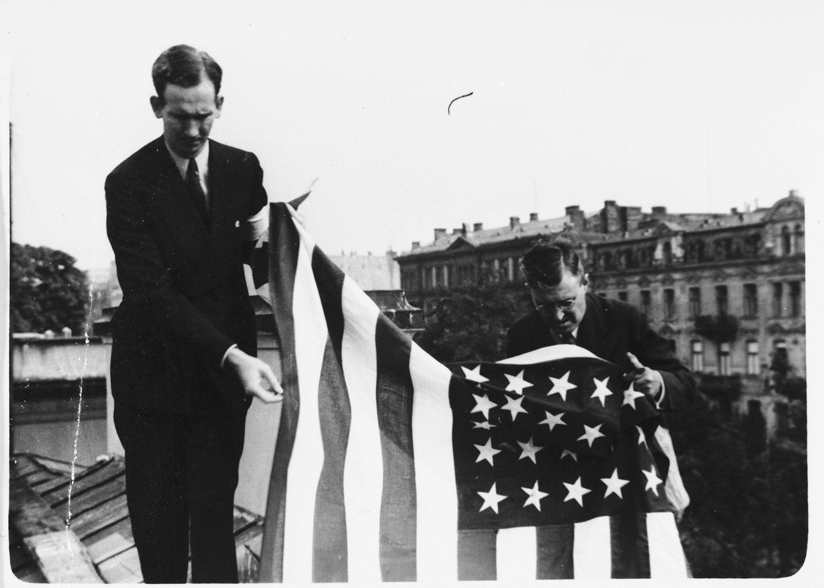 Two American consular staff members hang an American flag from the roof of the embassy in Warsaw in anticipation of the arrival of German troops into the capital.  

Photographer Julien Bryan describes the scene:  "On September 12 [1939] word came over the radio that the German army was entering the city.  Out came all the available American flags.  The largest - ten by fifteen feet - was carried to the roof.  The other flags we suspended in front of the embassy and also at the rear, believing the German troops might approach from that direction.  Finally we placed one small flag over the dugout in the garden.  But the Germans did not come that day, nor did they come while we were in Warsaw." [Source: Bryan, Julien.  "Warsaw 1939 Siege; 1959 Warsaw Revisited.  Warsaw, Polonia, 1959, p. 31.]