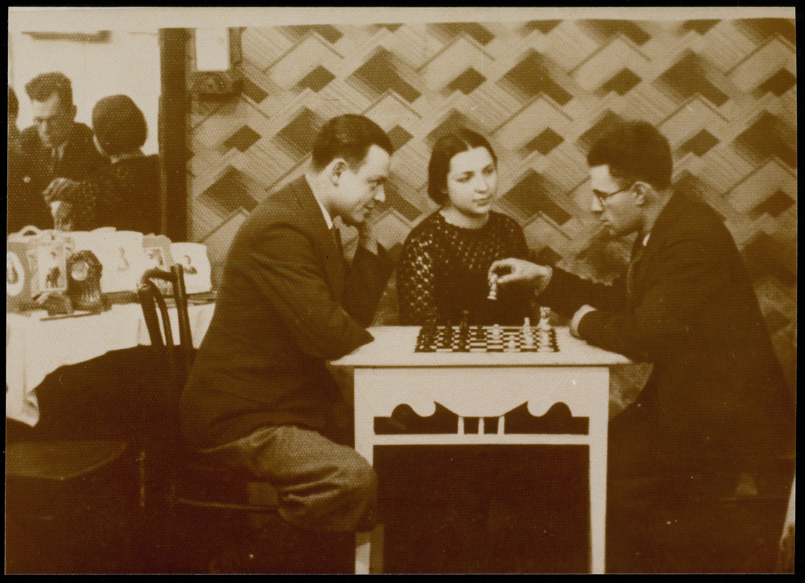 Two men play chess while Malka Matikanski looks on.

Malka Matikanski sitting in the center and Velvke Kaganowicz is on the right.  The man on the left is unidentified.  The photograph was taken in the photographer's house on Vilna Street.

Malka Matikanski  immigrated to Palestine.  Velvke Kaganowicz, his wife and children were murdered by the Germans during the September 1941 killing action in Eisiskes.