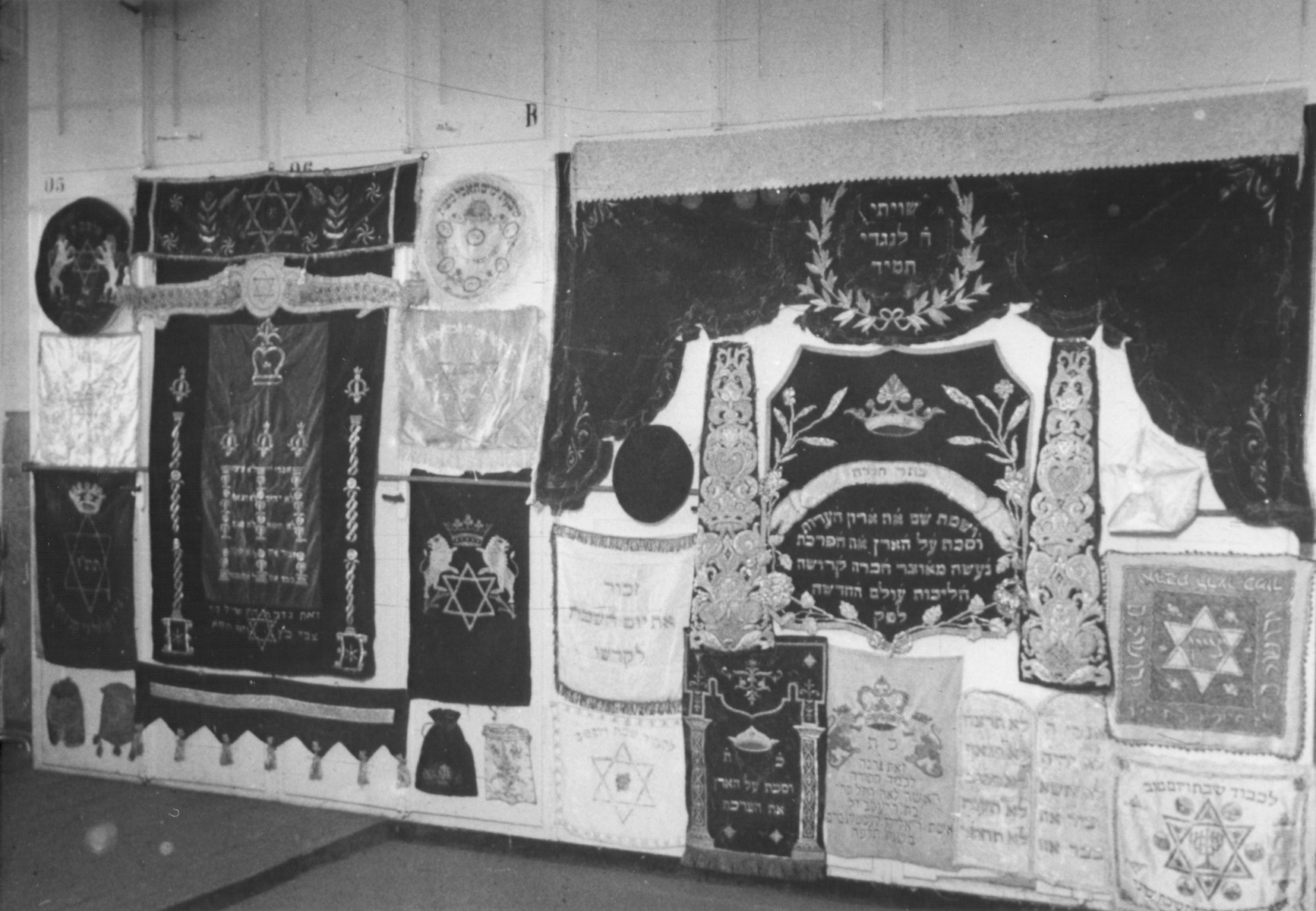 Display of ritual synagogue textiles confiscated by the Nazis.

The Torah curtain, bottom row, third from the right, is now part of the collection of the Jewish Museum of Westphali. They acquired the curtain from art trade "Pieces of Time," London in 1987 and provided the caption and following translation:

"The historically valuable Torah curtain dates from 1925 and it is white for the high religious occasions. The donors were Leah Kestenbaum (1864-1949) and her husband Reb Elijahu Kestenbaum (1862-1936) from Leipzig. The family emigrated in the 1930s to New York, where their Descendants still live today. The curtain has the following embroidery:

This is a gift in honor of the torah Mrs. Leah Rachel, may she live/long she shall live, Daughter of Reb Jakob, blessed memory (s..l: sichrono li-wrachah
The wife of Reb  Elijah (u) Kestenboim/Kestenbaum
In the year 1925"