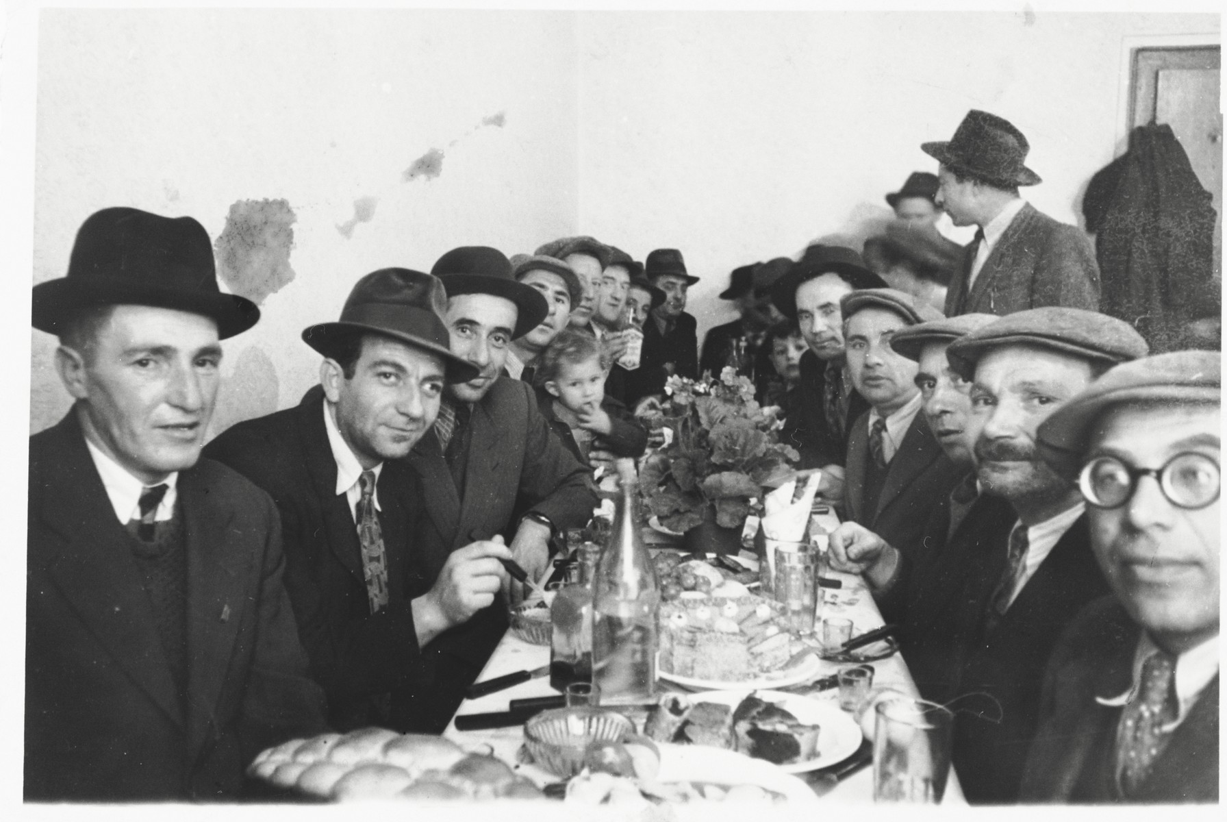 Jewish DPs are gathered around a table at a festive meal following the circumcision of Arele Werkel, the son of Taibel and Yehiel Greenberg at the Ebelsberg displaced persons camp near Linz, Austria.

Among those pictured are Itzhak Gendelman (standing at the upper right).