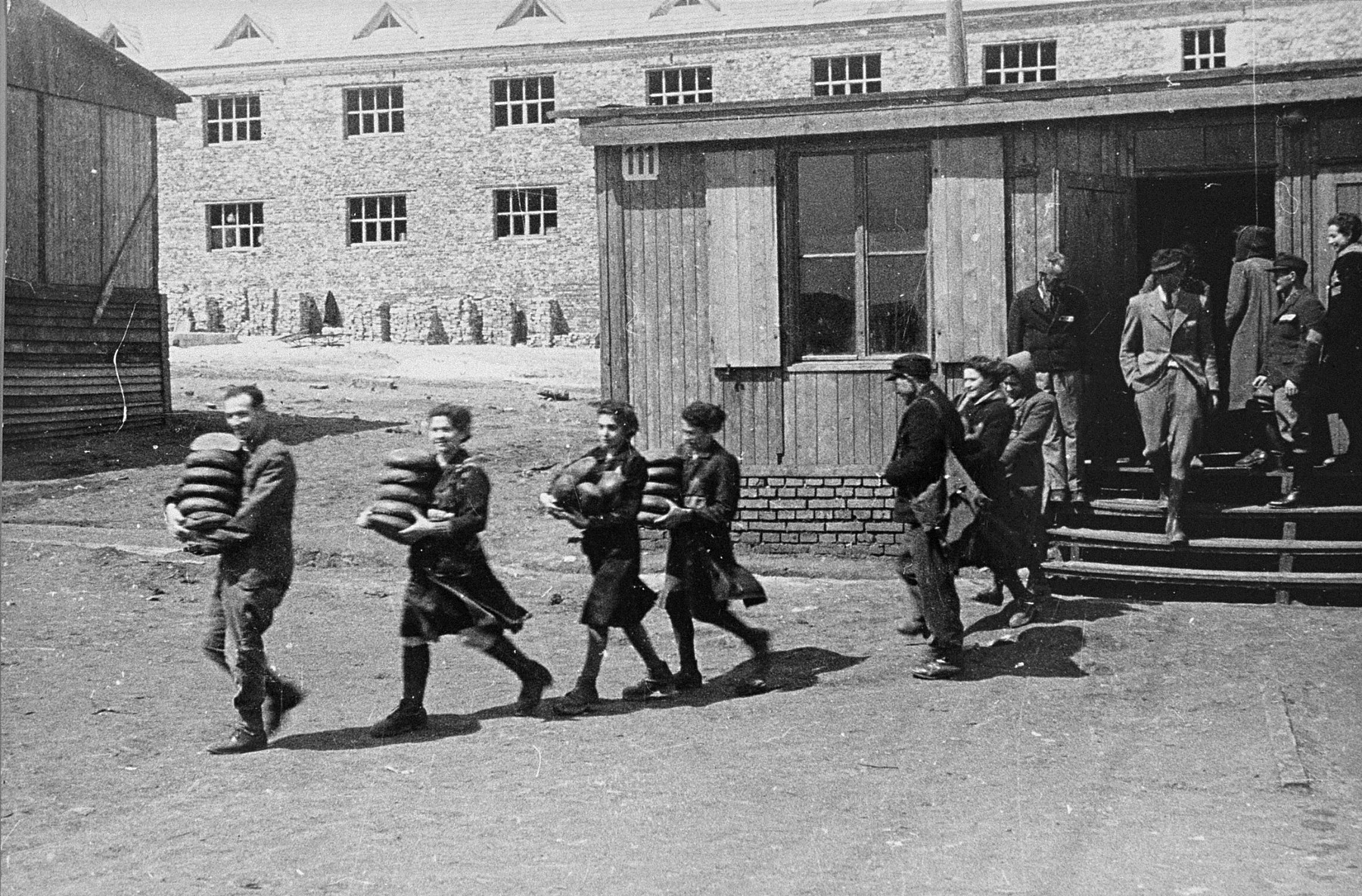 Jewish prisoners carry loaves of bread from a storehouse to the Madritch factory.  

The Madritch factory utilized concentration camp labor to produce uniforms for the German army.  The man to the right with the armband is likely a civilian supervisor.