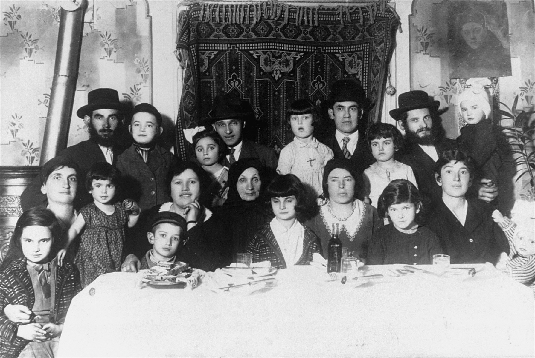 Group portrait of the family of Elizabeth Ehrlich seated around the family table in Mukachevo. 

Among those pictured is Elizabeth Ehrlich (later Roth) standing in the middle of the back row in a light dress, and Rella Ehrlich (front row, second from the right).  Elizabeth was born March 26, 1926 in Mukachevo, and in 1944 was confined to the ghetto there before being deported with her family to Auschwitz.  She was later transferred to a camp in Bydogszcz, Poland, and from there to the Stutthof concentration camp.